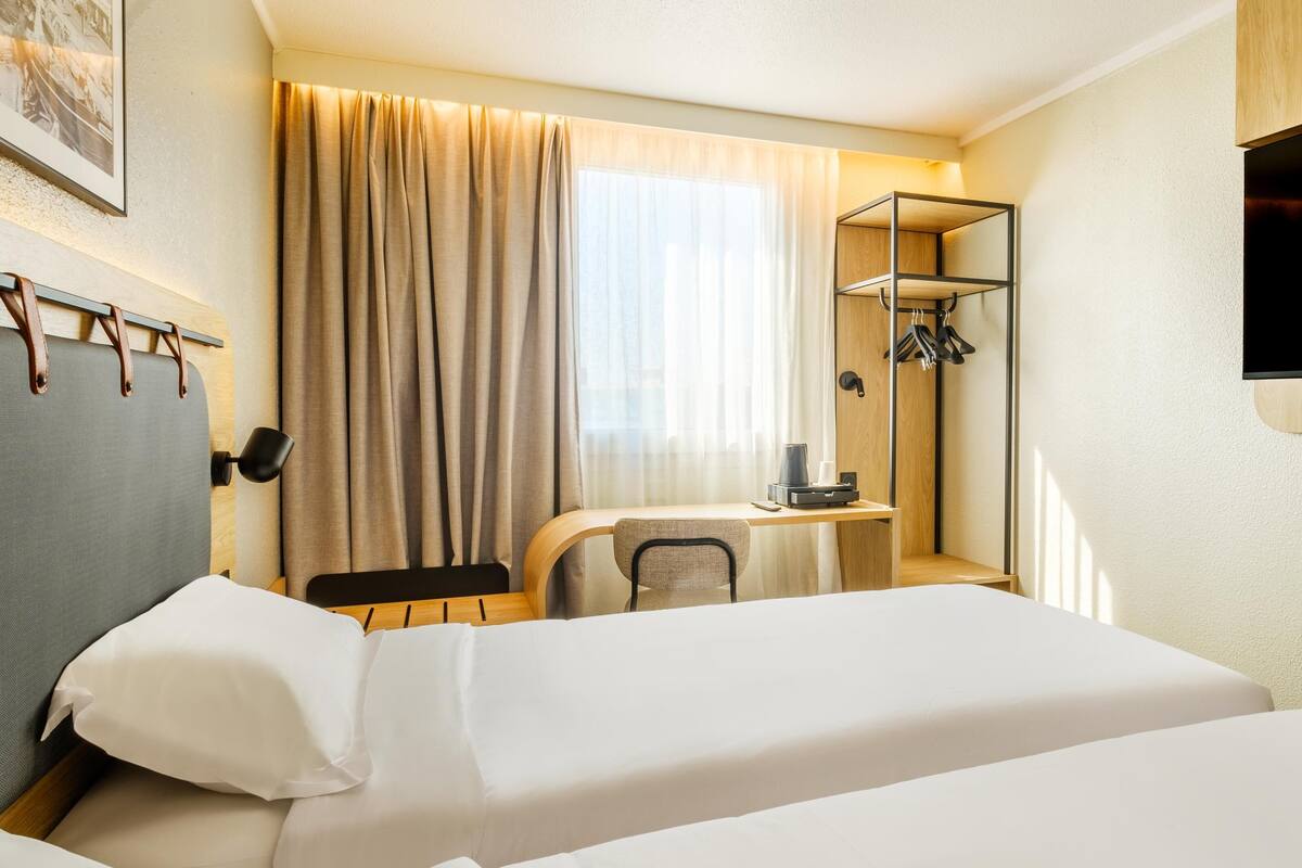 A hotel room with a bed, a desk with a chair, a TV mounted on the wall, and a coat rack.