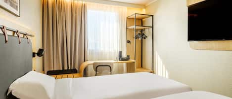 A hotel room with a bed, a desk with a chair, a TV mounted on the wall, and a coat rack.