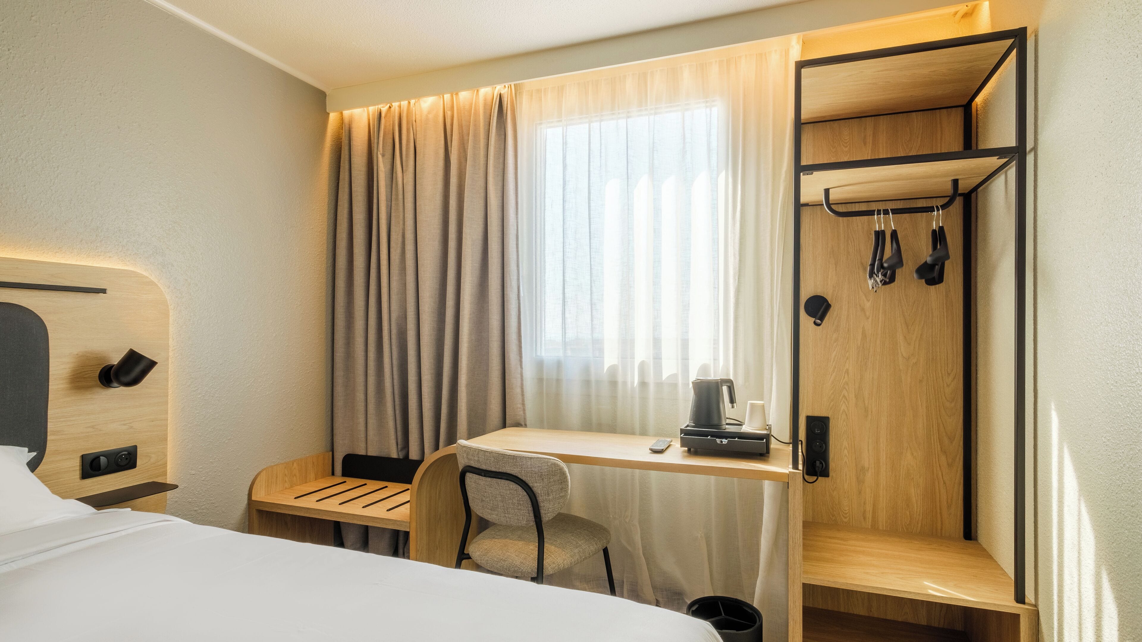 Ein kleines Hotelzimmer mit einem Bett, einem Schreibtisch mit Stuhl, einer Kaffeemaschine und einem Kleiderhaken.