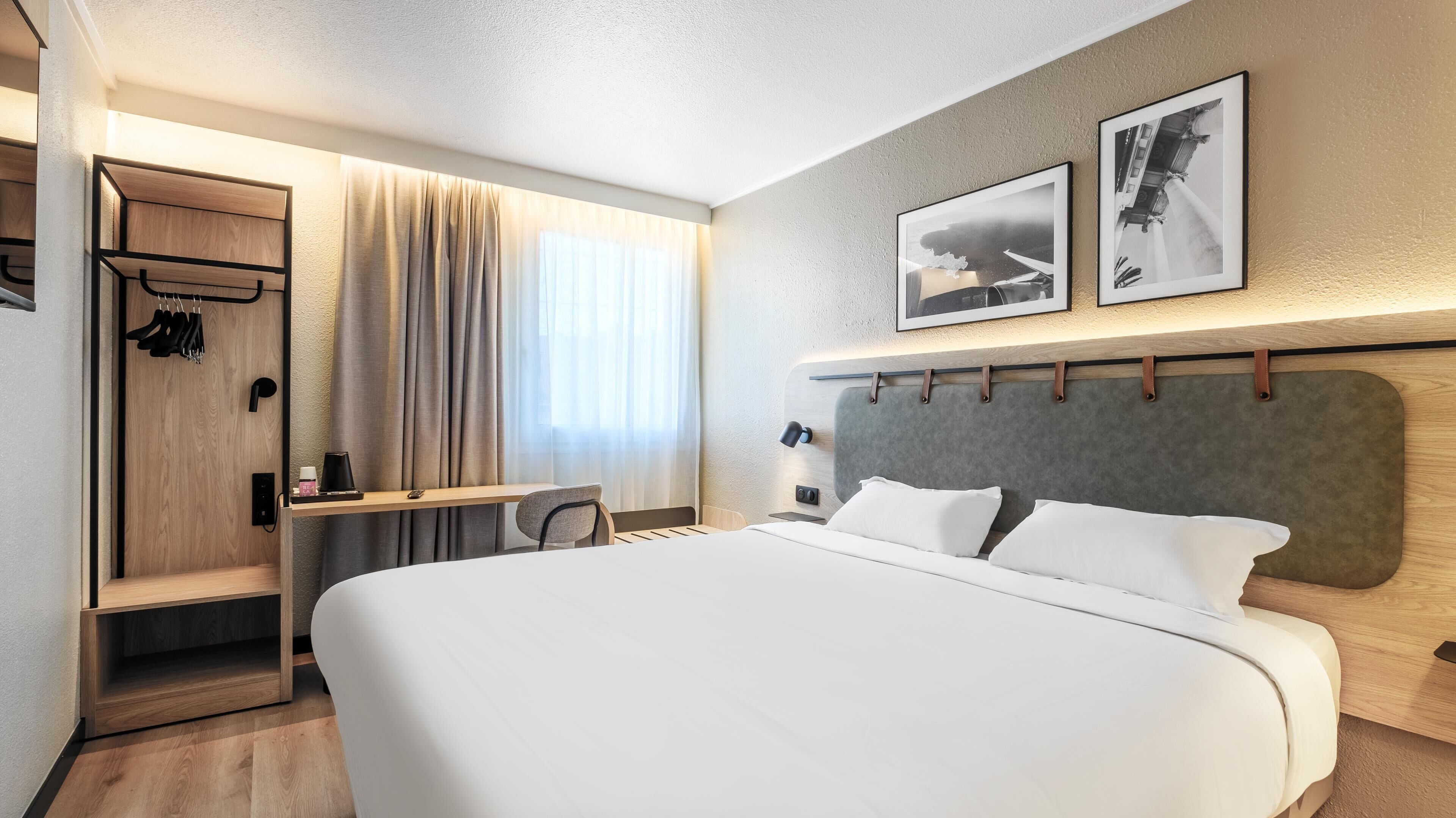 Ein modernes Hotelzimmer mit einem großen Bett, einem Schreibtisch mit Stuhl, einem Kleiderschrank und zwei gerahmten Bildern an der Wand.