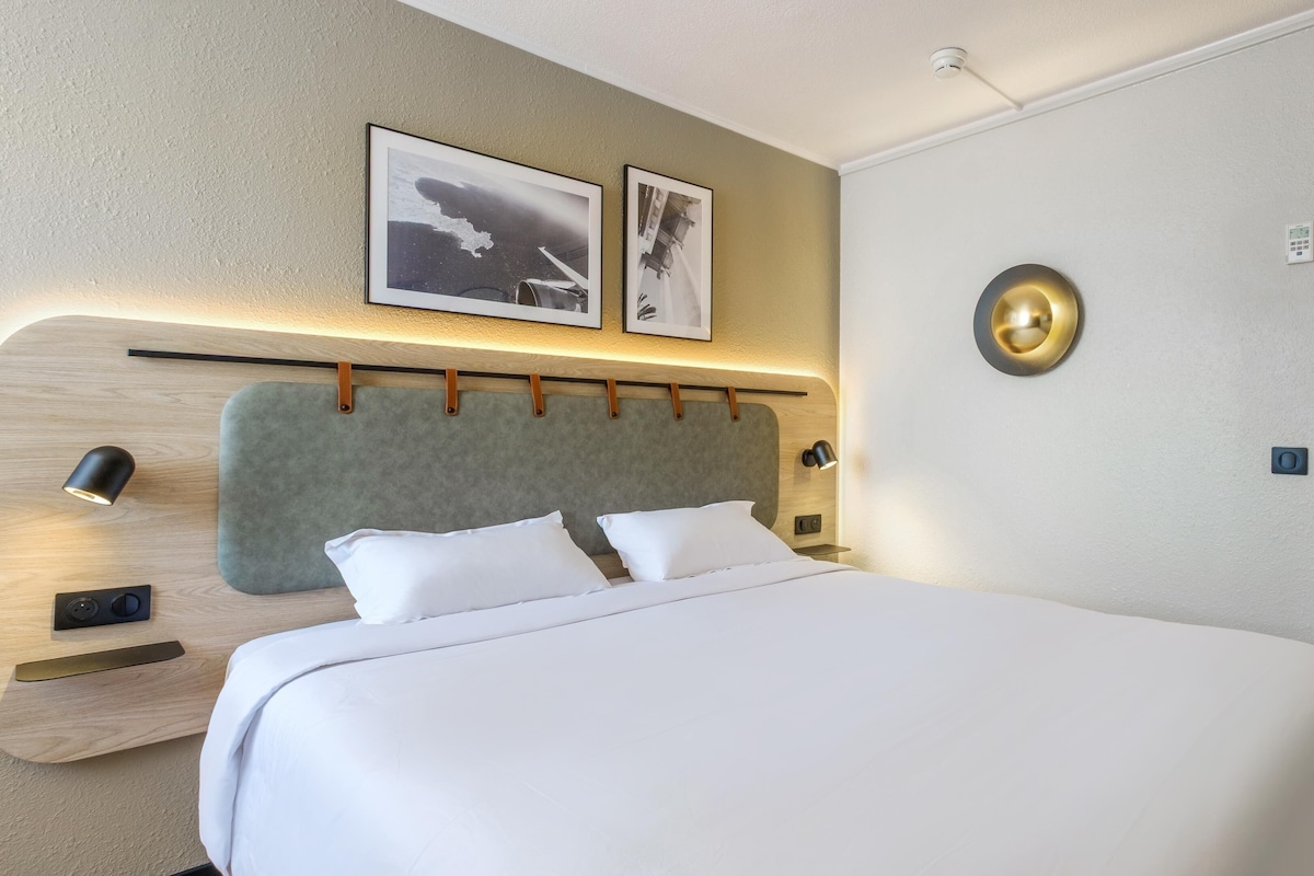 A hotel room with a large bed, two wall-mounted picture frames, a bedside table with a lamp, and a round wall clock.