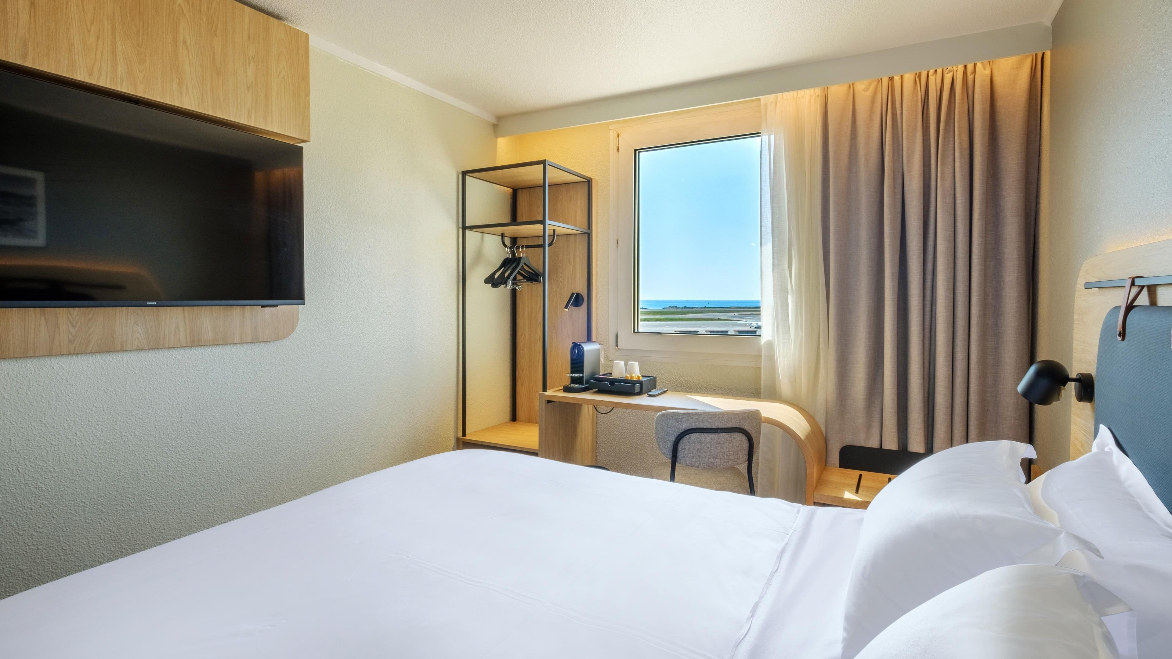 A hotel room with a bed, a desk with a chair, a TV mounted on the wall, a window with curtains, and a view of the beach.