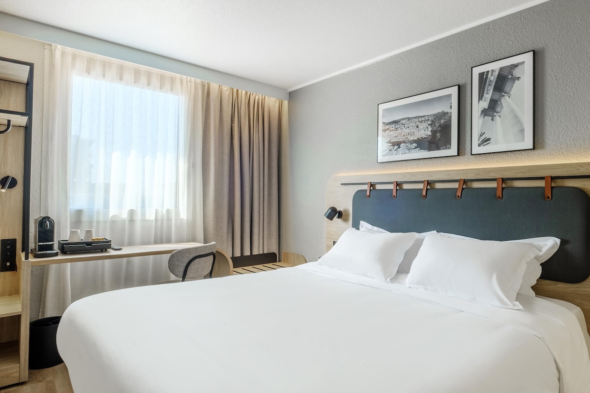 A hotel room with a large bed, a desk, a chair, and a window with curtains. There are two framed pictures on the wall above the bed.
