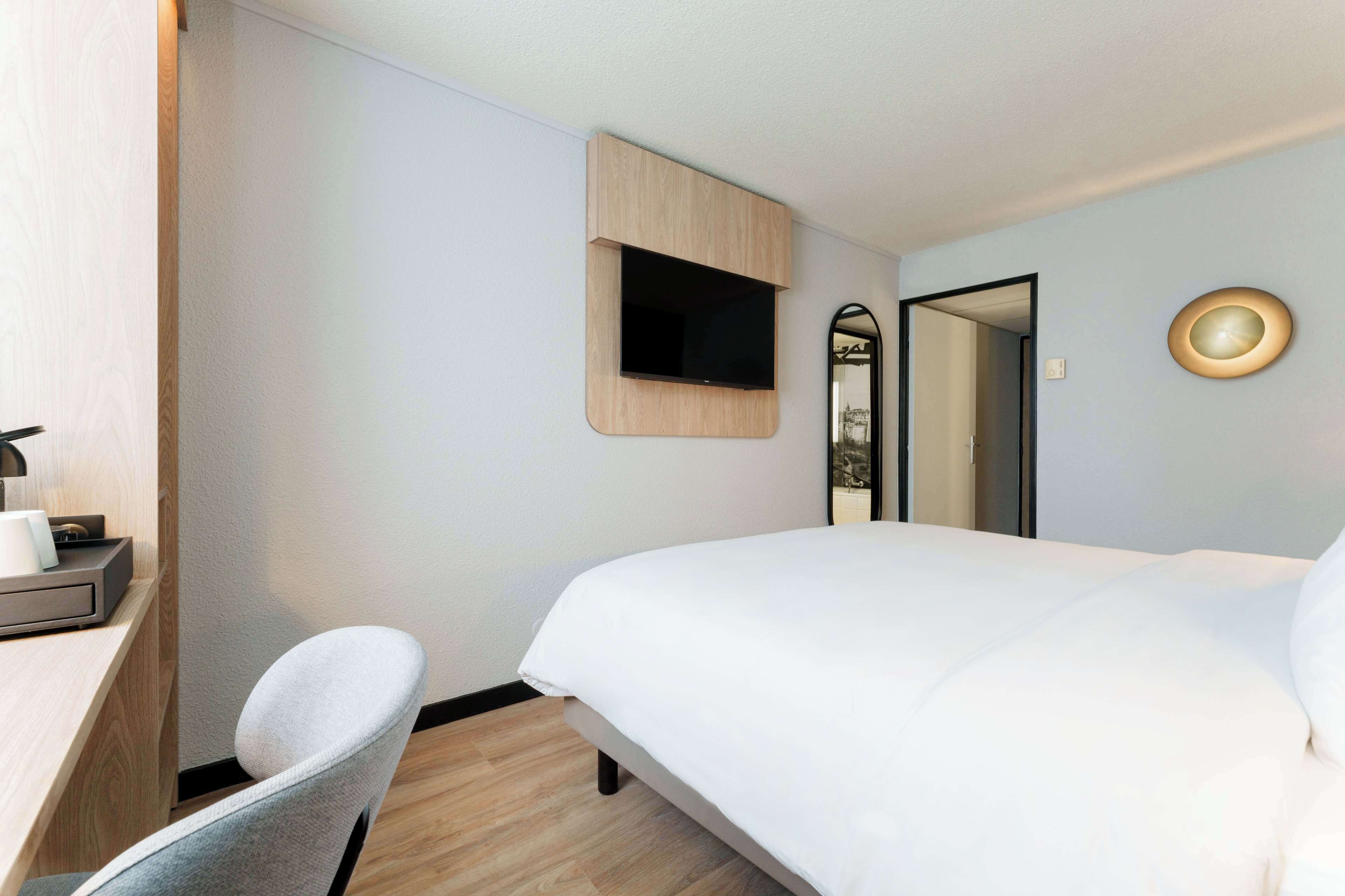 A hotel room with a bed, a desk with a chair, a TV mounted on the wall, and a mirror.
