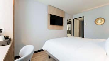 A hotel room with a bed, a desk with a chair, a TV mounted on the wall, and a mirror.