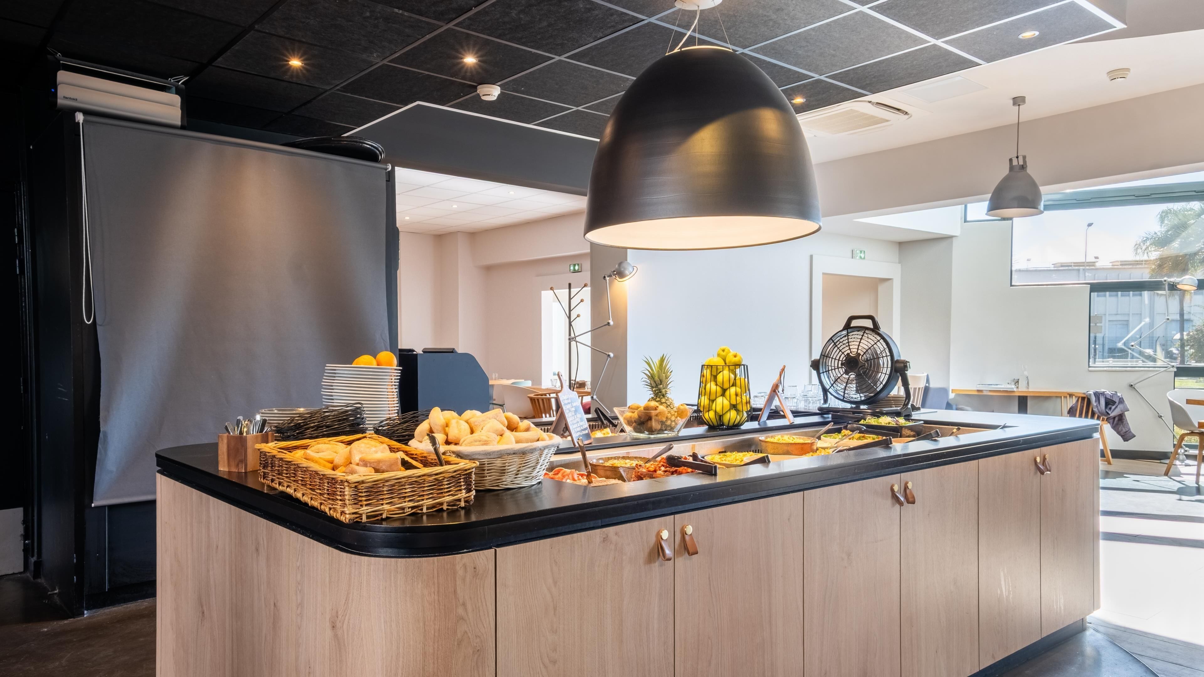 A modern kitchen with a central island stocked with breakfast items, a large black pendant light, and a view of the outdoors.