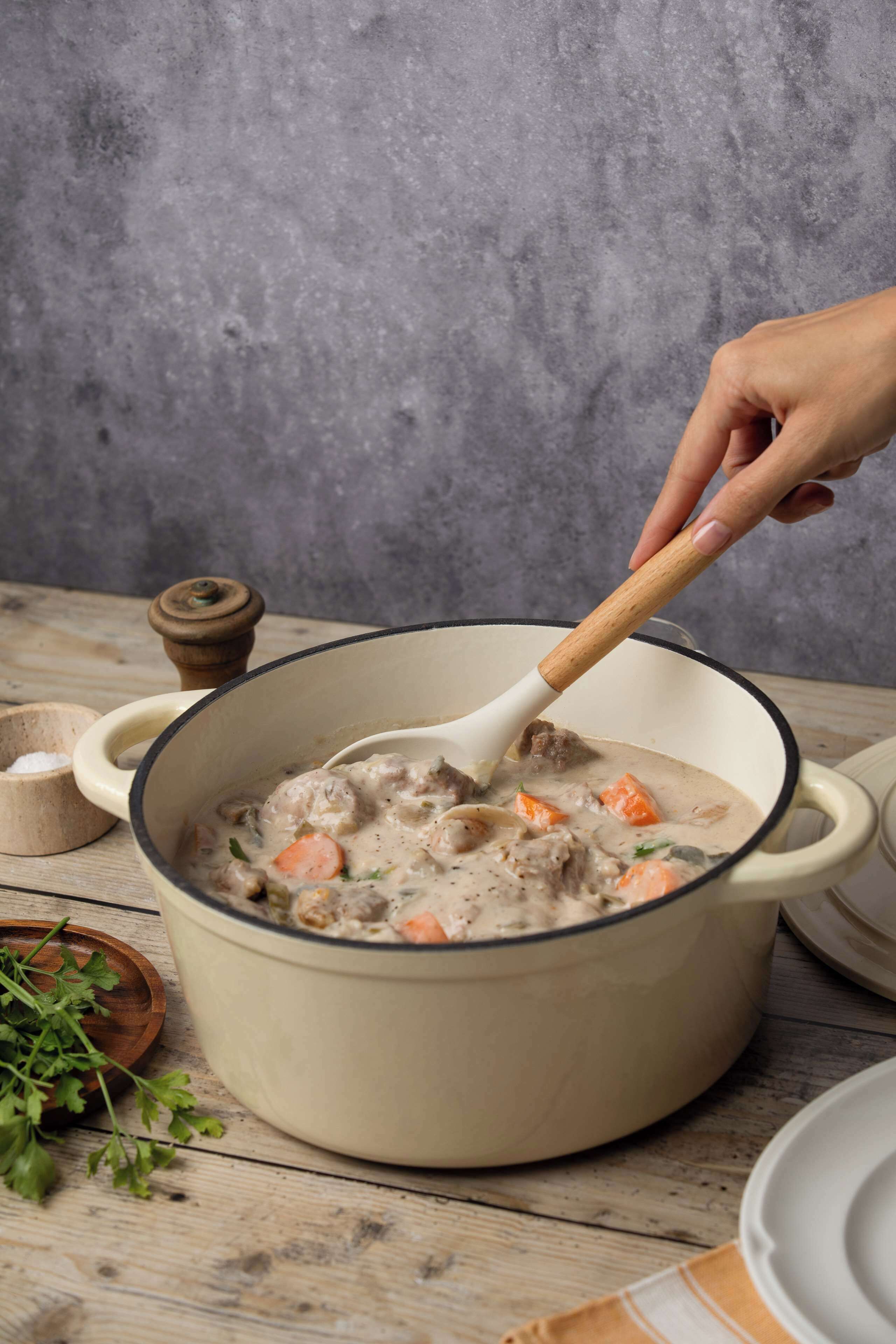 A large pot of stew with a wooden spoon, surrounded by bowls and a pepper grinder on a wooden table.