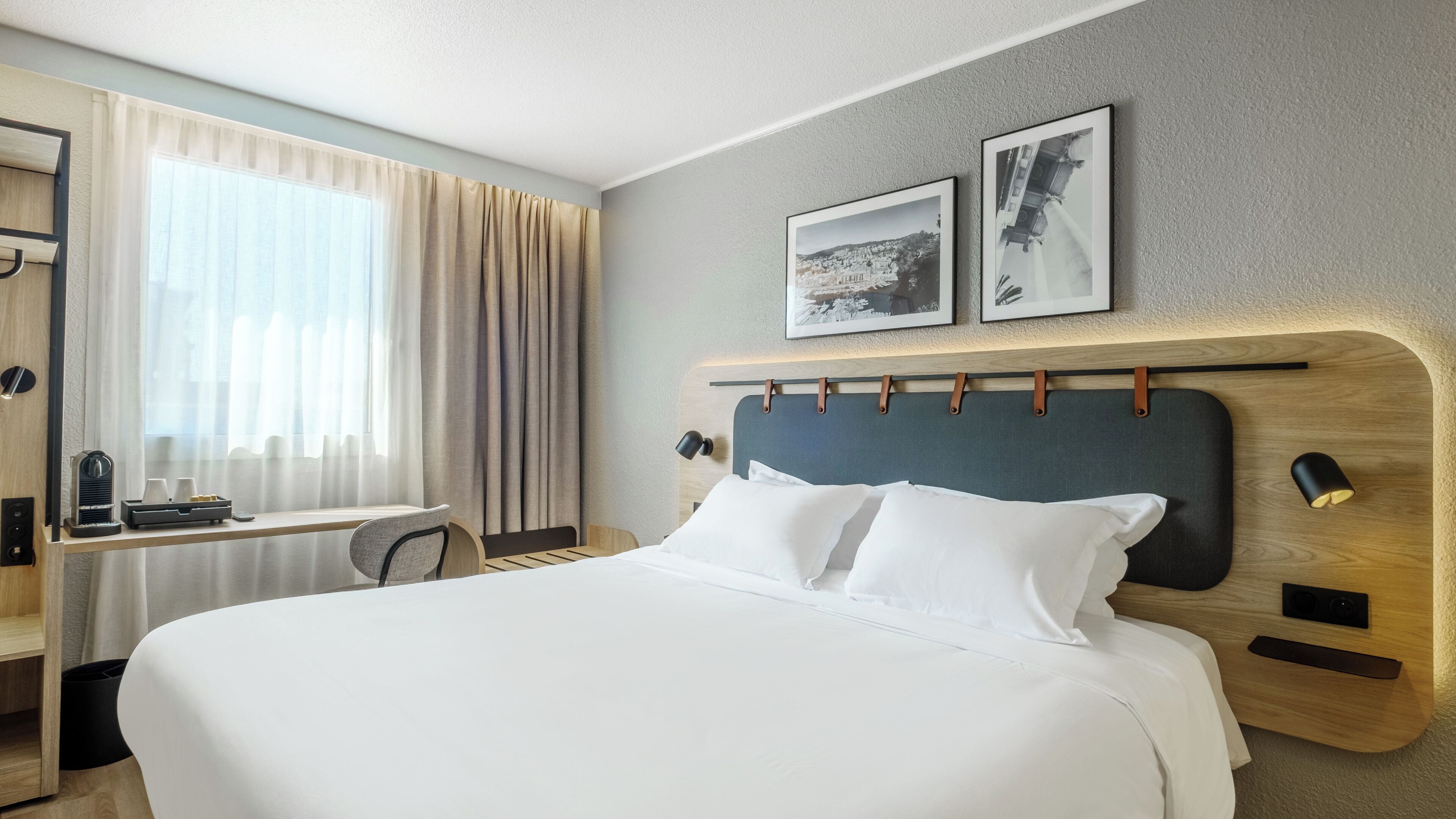 A hotel room with a large bed, a desk, a chair, and a window with curtains. There are two framed pictures on the wall above the bed.