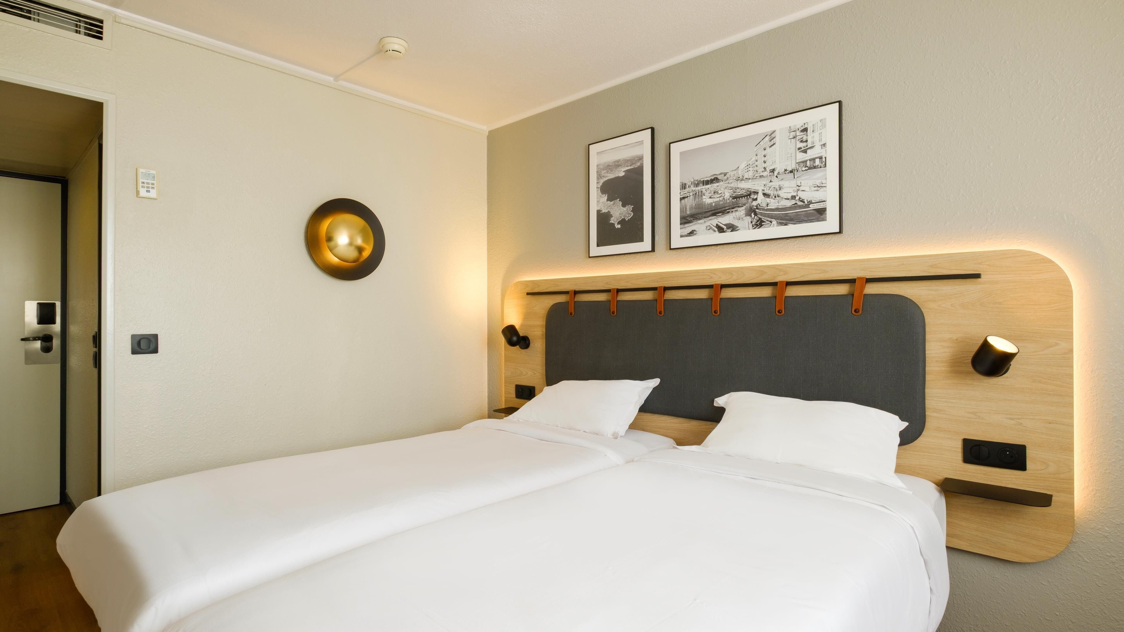 A hotel room with a double bed, a wooden headboard, two framed pictures on the wall, and a round wall light.
