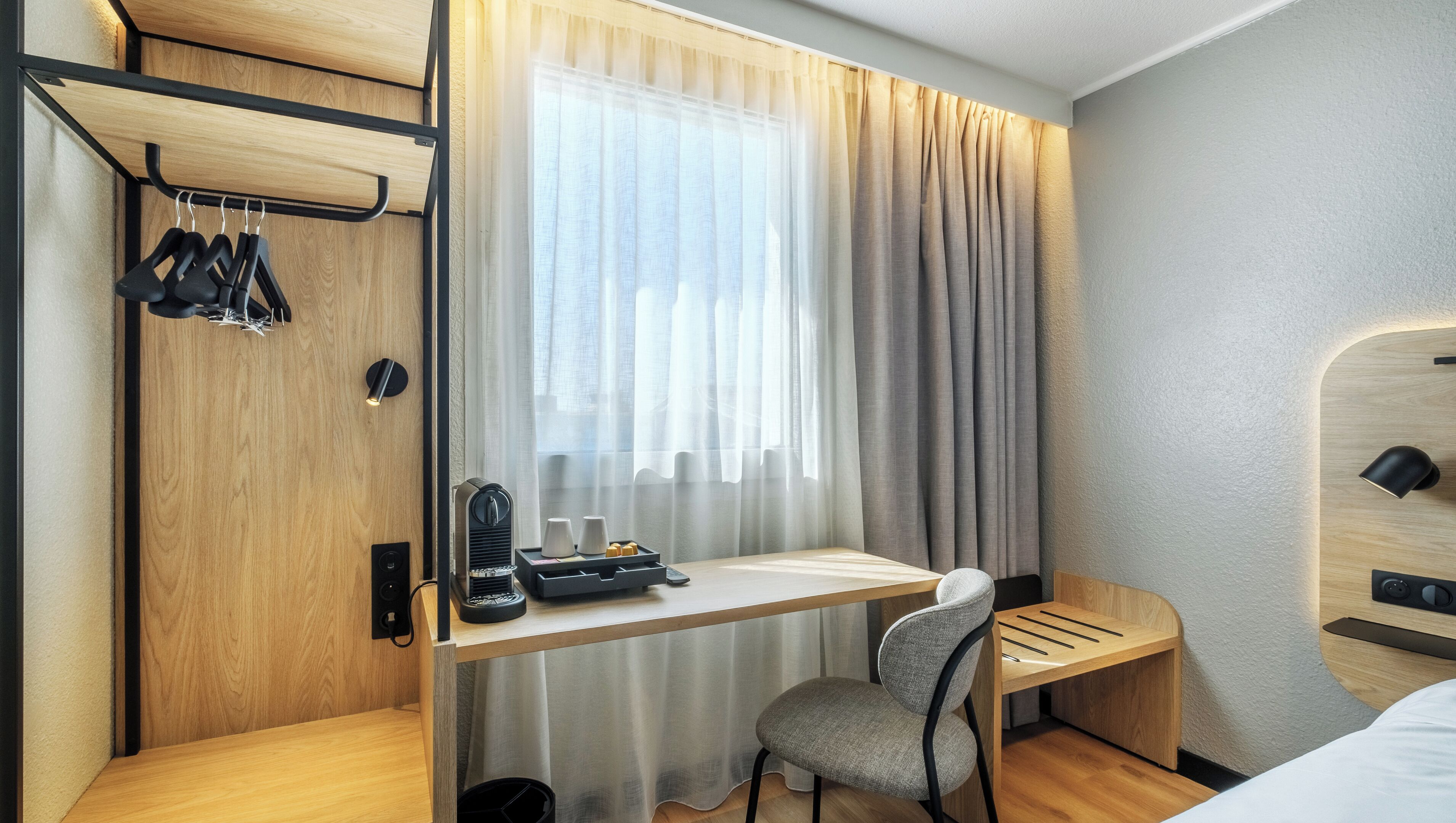 A compact hotel room with a wooden desk, a chair, a bed, a wardrobe with hanging clothes, and a window with curtains.