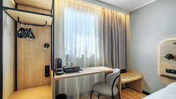 Een kleine hotelkamer met een houten bureau, een stoel, een bed, een kledingkast met daarin hangende kleding en een raam met gordijnen.