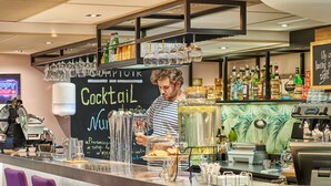 Zona de bar con un cantinero preparando bebidas, estantes con botellas y vasos, y un menú en la pizarra.