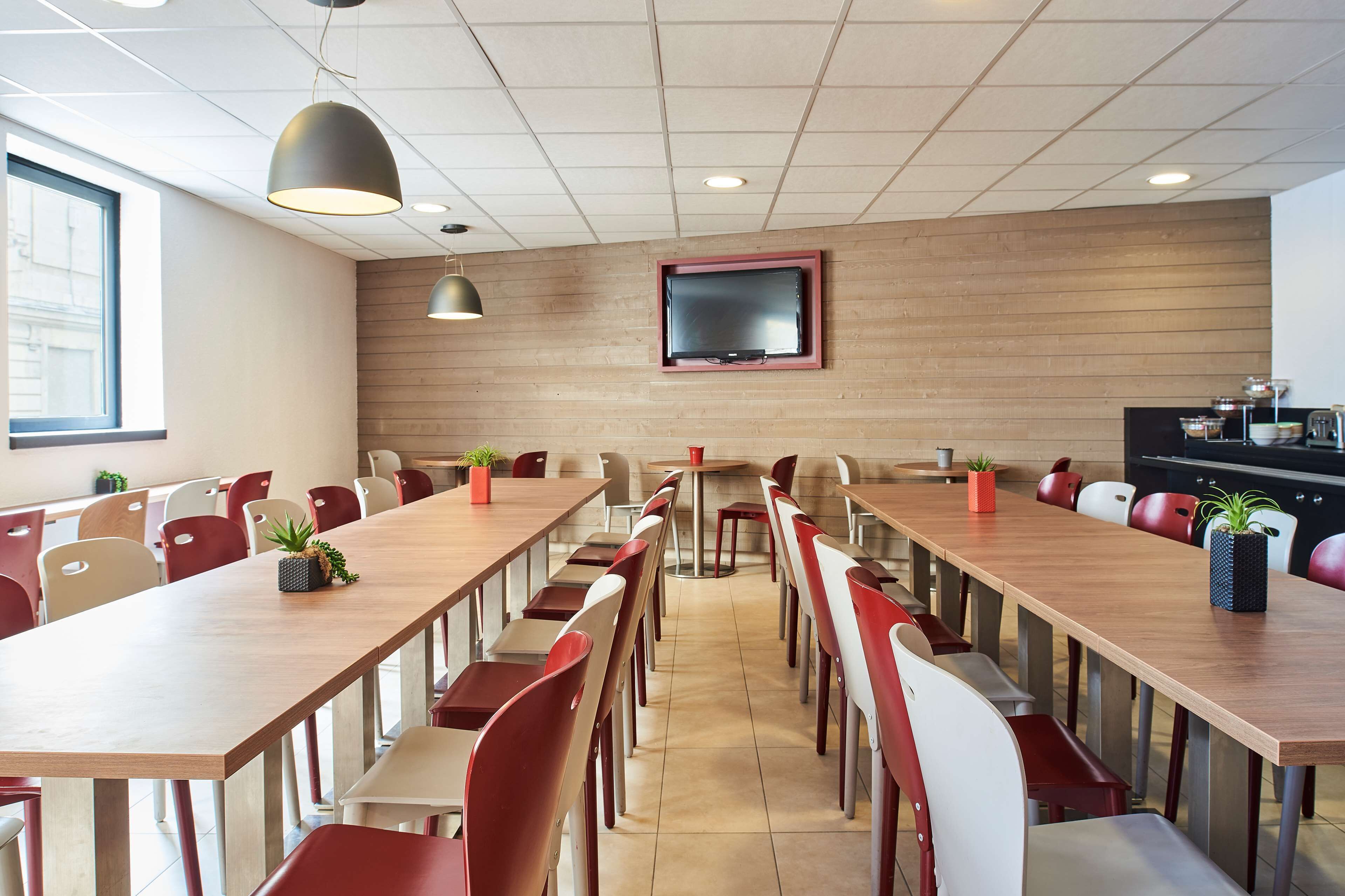 A conference room with long tables, red and white chairs, potted plants, and a television on the wall.
