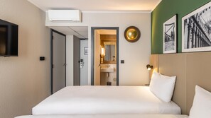 Habitación de hotel con cama, televisión, equipo de aire acondicionado, baño con lavamanos y espejo, y cuadros en la pared.