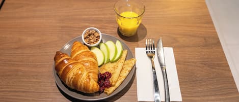 Un plato con croissants, rebanadas de manzana y un cuenco pequeño de granola, acompañado de un vaso de jugo de naranja y cubiertos sobre una mesa de madera.