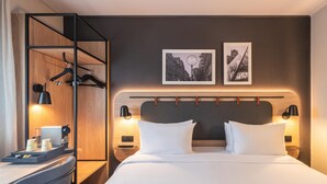 Habitación de hotel con una cama, lámparas de noche, un armario de madera, un escritorio con lámpara y dos cuadros en la pared.