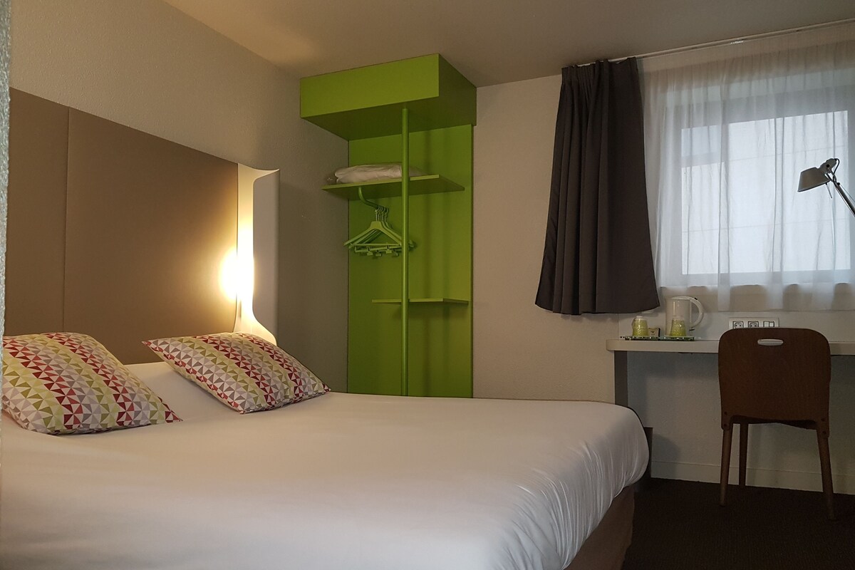A hotel room with a bed, a desk, a chair, a green shelving unit, and a window with curtains.