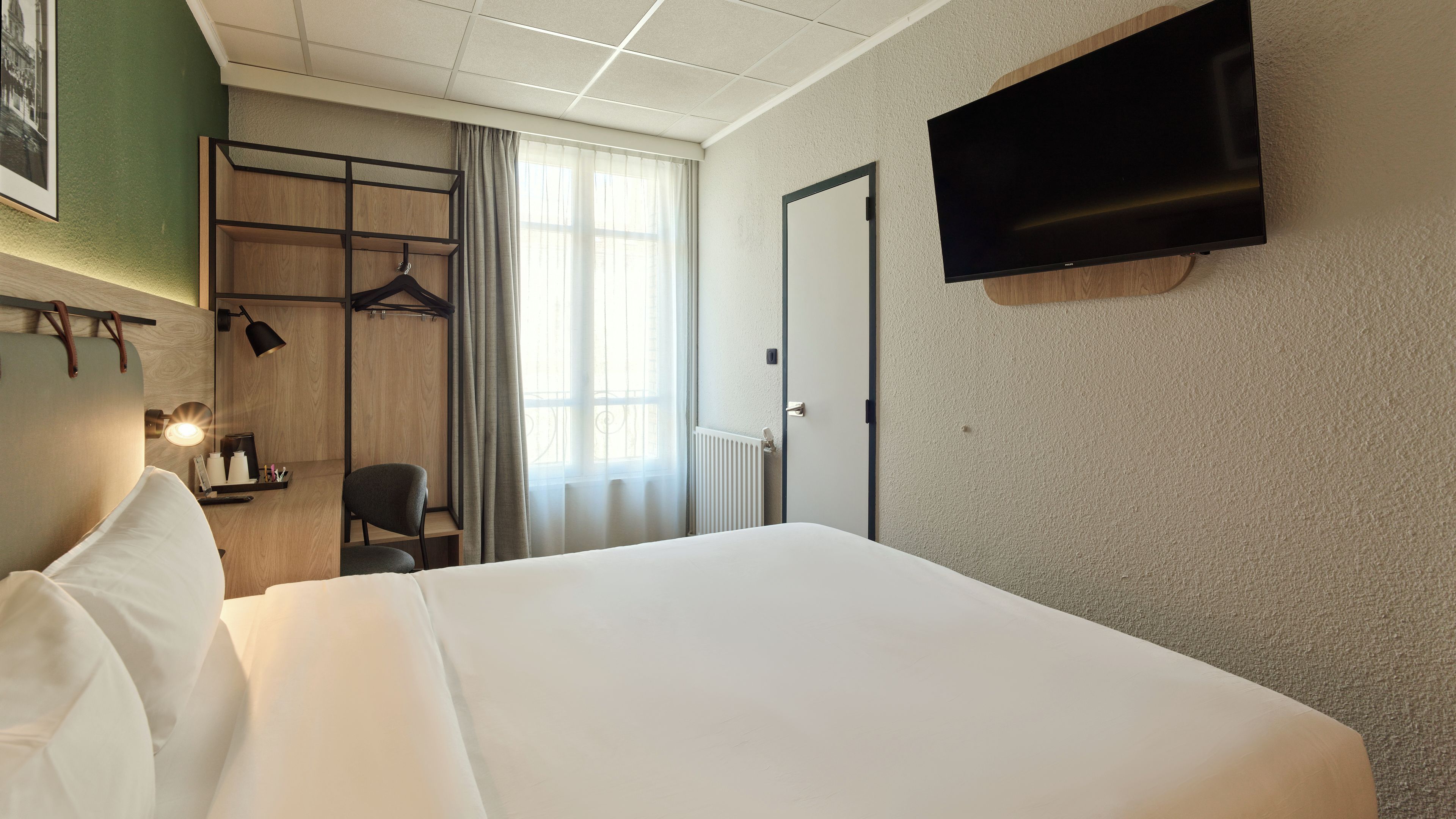 Habitación de hotel con cama, escritorio con silla, televisor montado en la pared y ventana con cortinas.