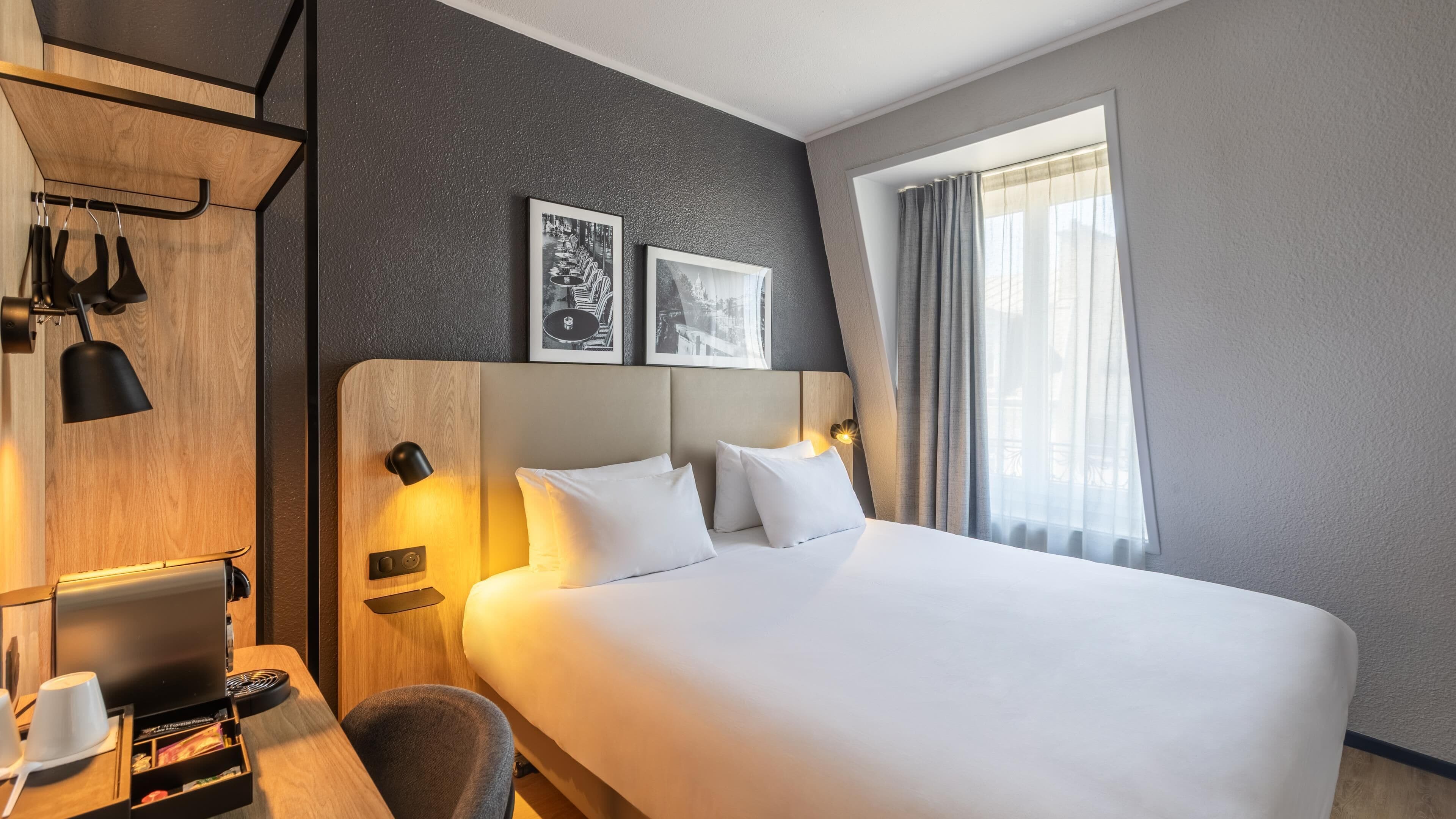 A hotel room with a bed, a desk with a computer, a chair, and a coffee machine. There are two framed pictures on the wall above the bed.