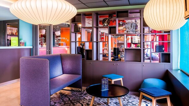 Un espacio lounge moderno con un sofá, una mesita redonda y dos sillones. Hay estanterías con diversos objetos y un ventanal con vistas.