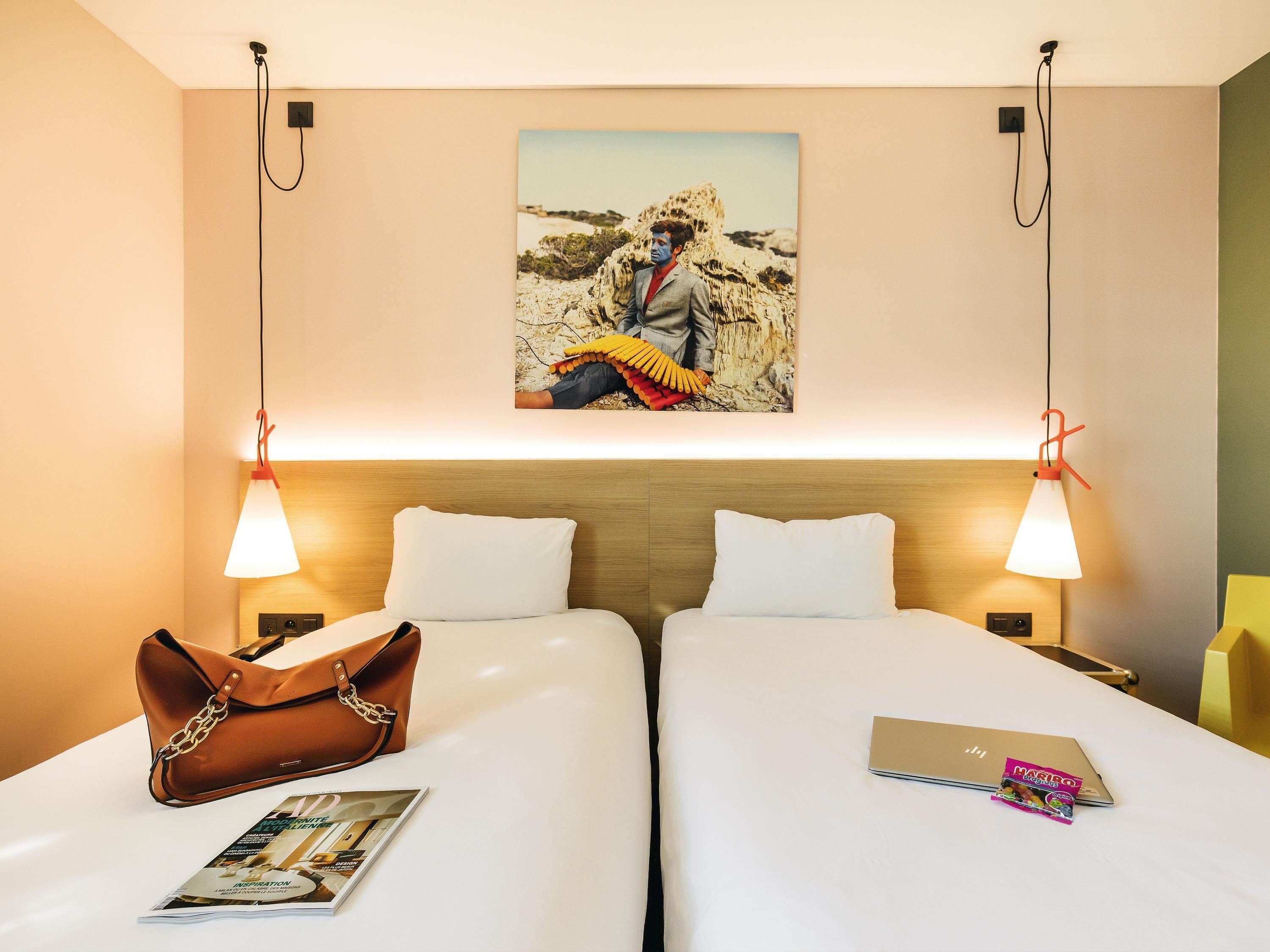 Una habitación de hotel con dos camas, un bolso marrón, una revista y un ordenador portátil sobre una de ellas.