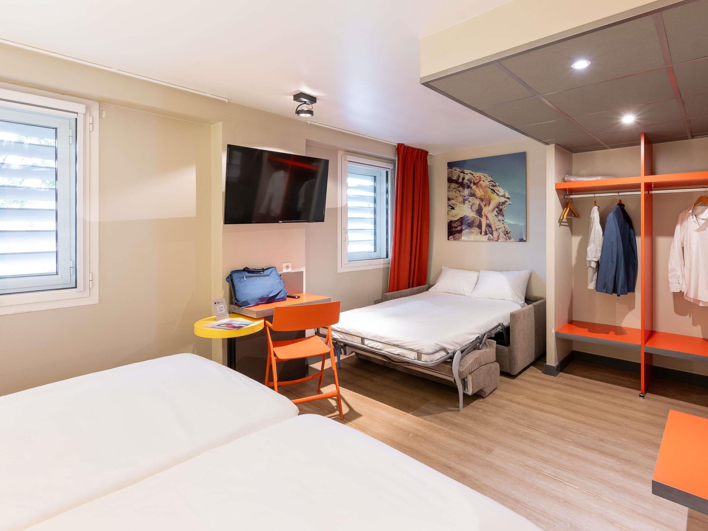 Een hotelkamer met een bed, een bureau met stoel, een televisie, een kledingkast en een raam met jaloezieën.