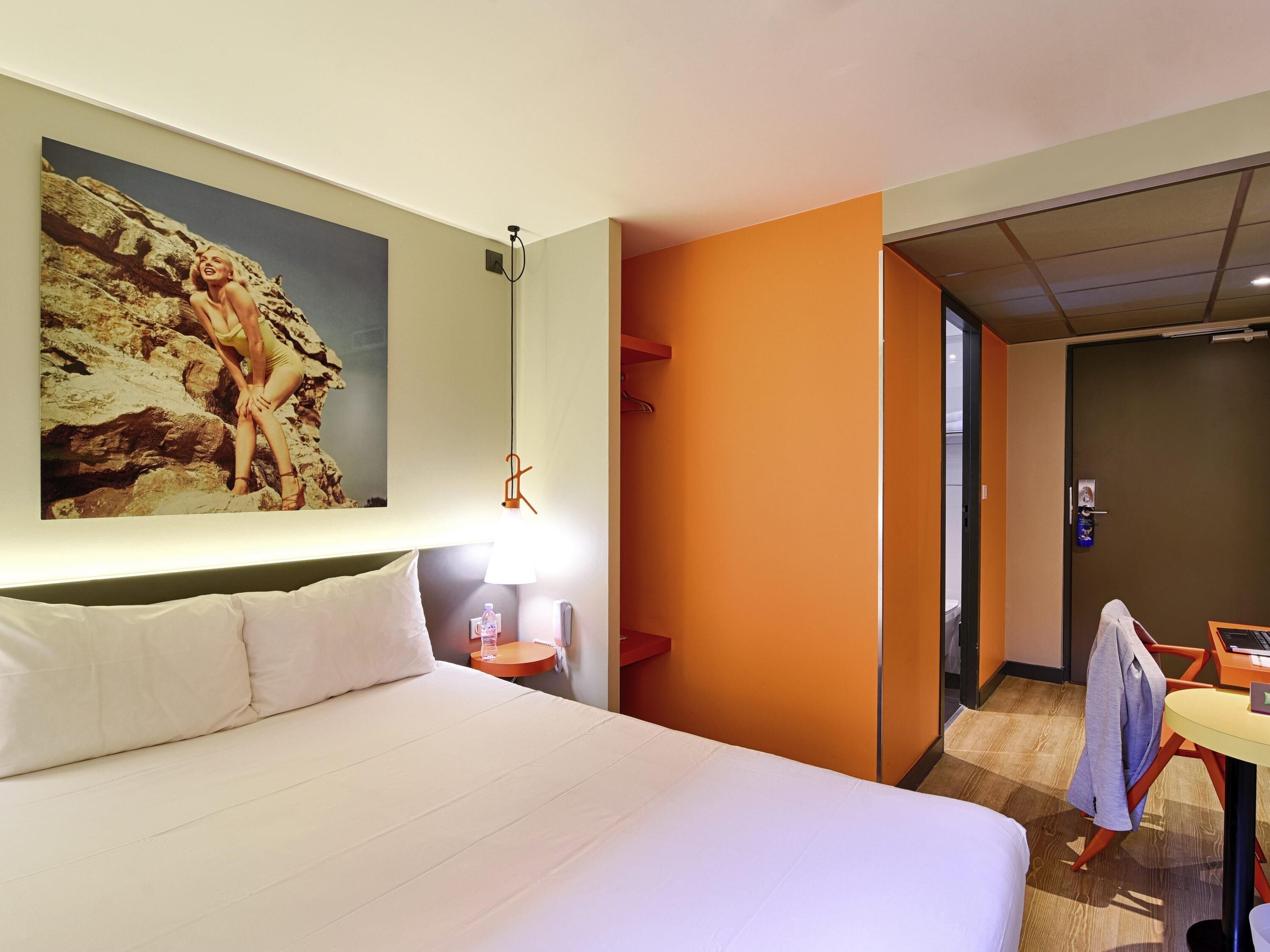A hotel room with a bed, a nightstand, a lamp, a painting, an orange accent wall, a desk with a chair, and a door.