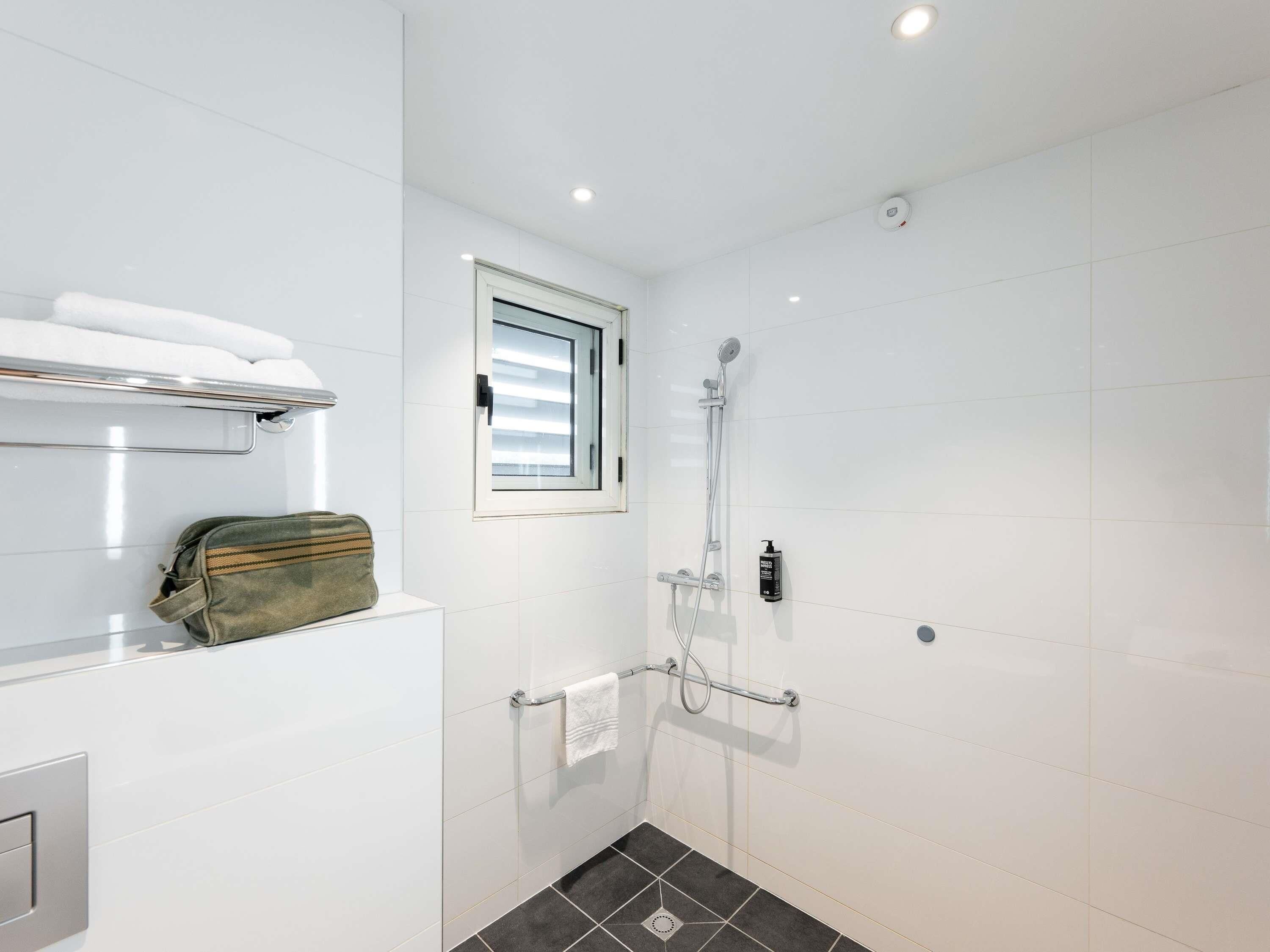 Un baño moderno con azulejos blancos, ducha con cabezal de ducha manual, toallero y ventana con persianas.