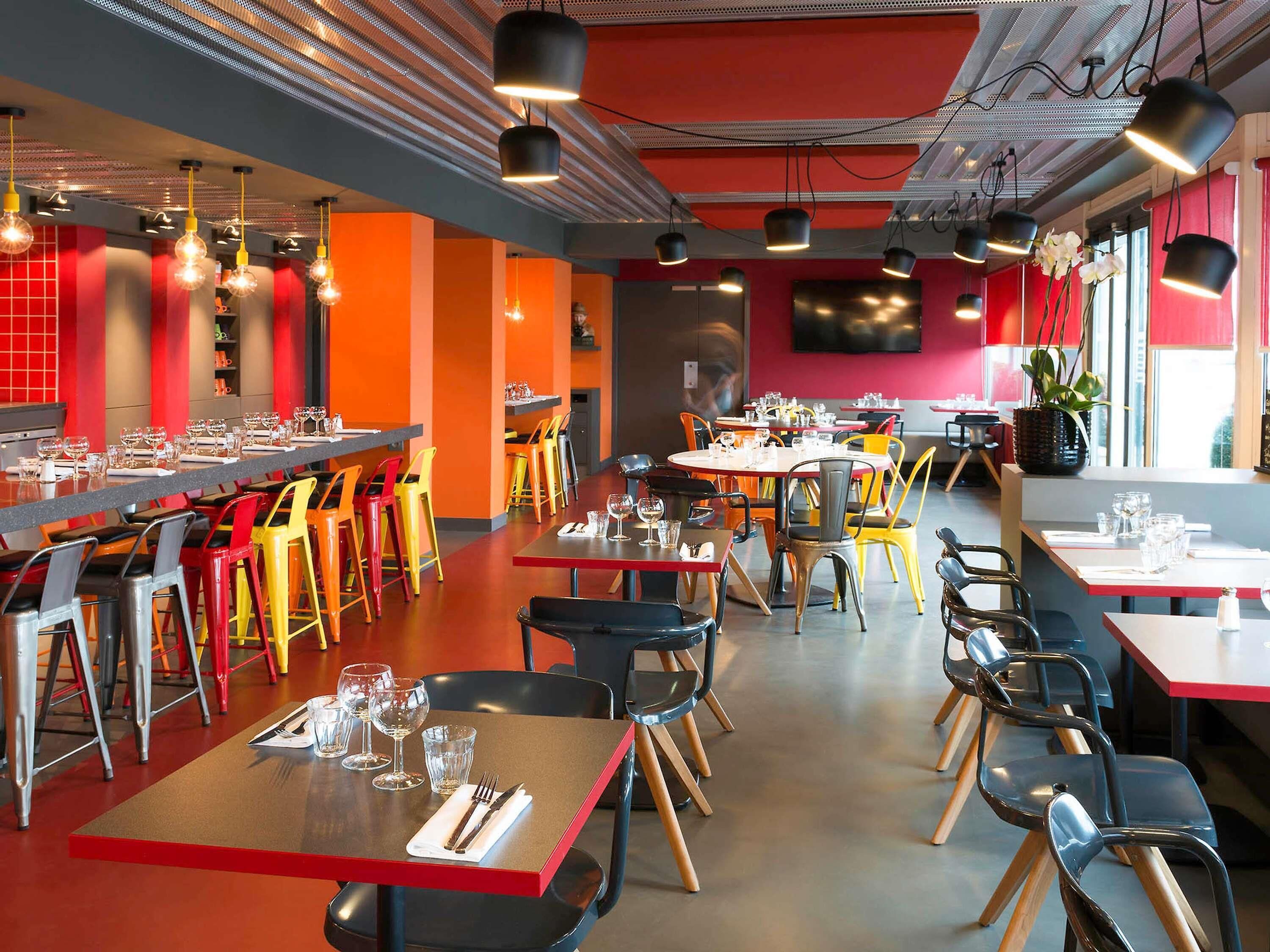 Een modern restaurantinterieur met rode en oranje wanden, meerdere eettafels met stoelen en hangende pendellampen.