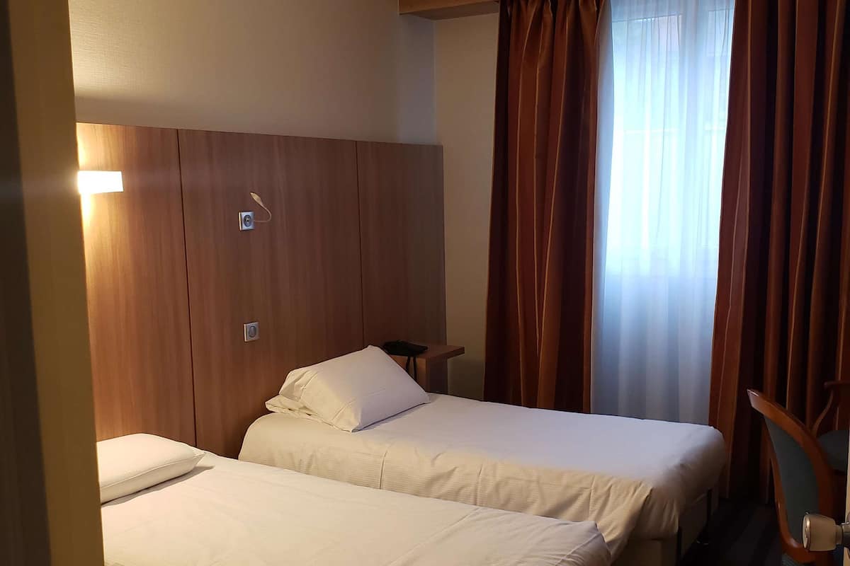 Habitación de hotel con dos camas, cabecero de madera, una mesita y una ventana con cortinas.