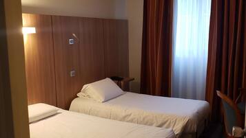 Habitación de hotel con dos camas, cabecero de madera, una mesita y una ventana con cortinas.