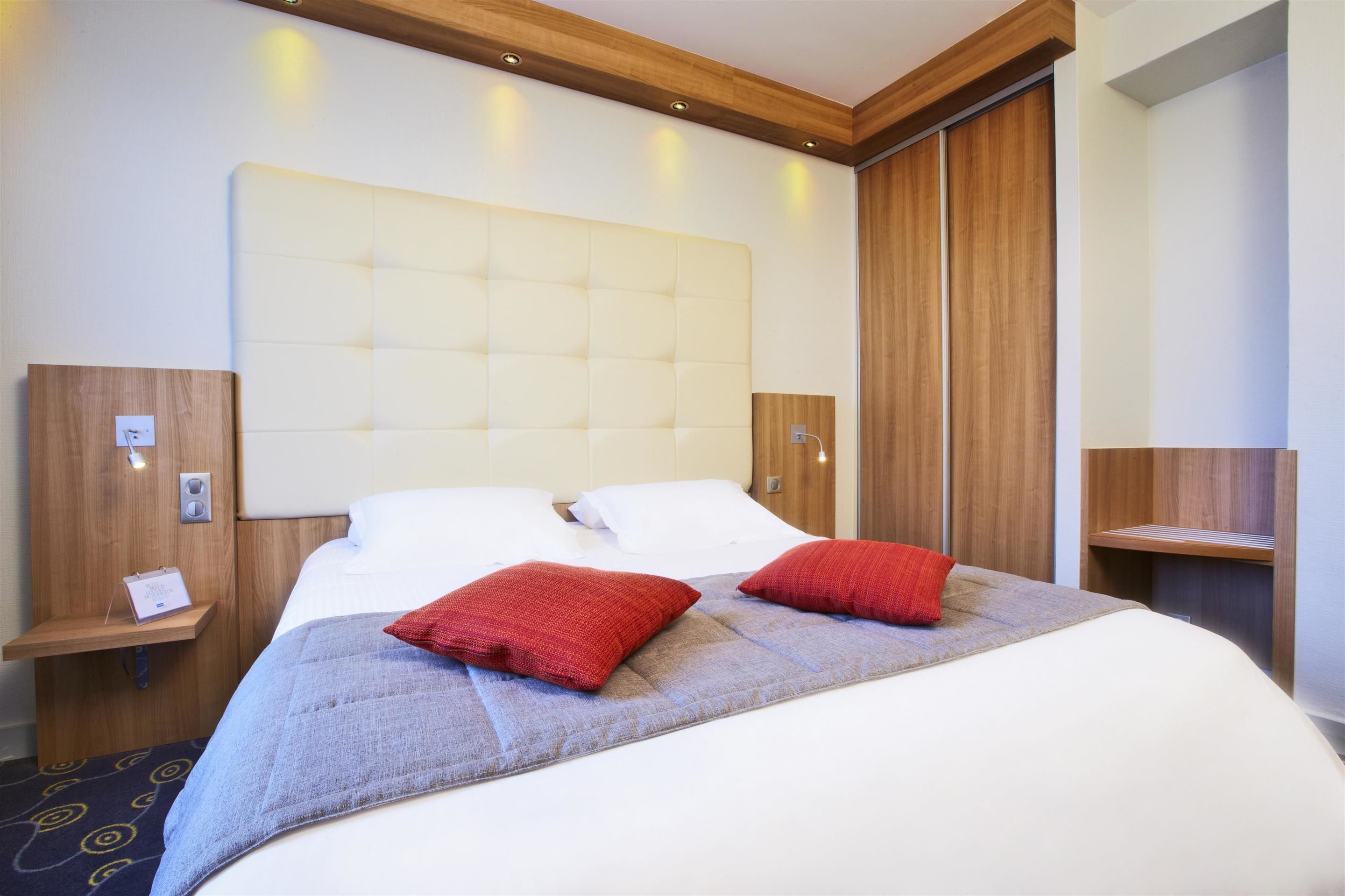 A hotel room with a large bed, two red pillows, a wooden headboard, and a nightstand with a phone.