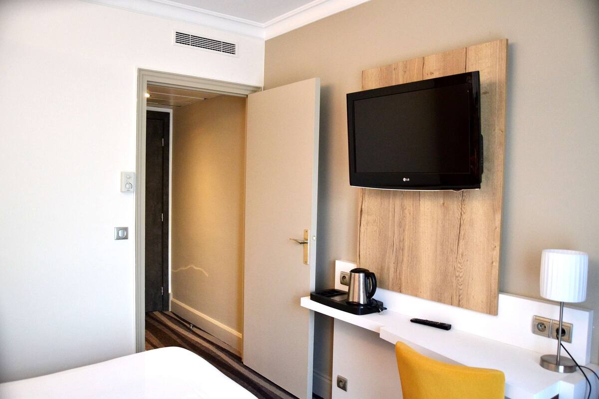 Habitación de hotel con cama, televisión montada en un panel de madera, cafetera, control remoto y lámpara en la mesita de noche.