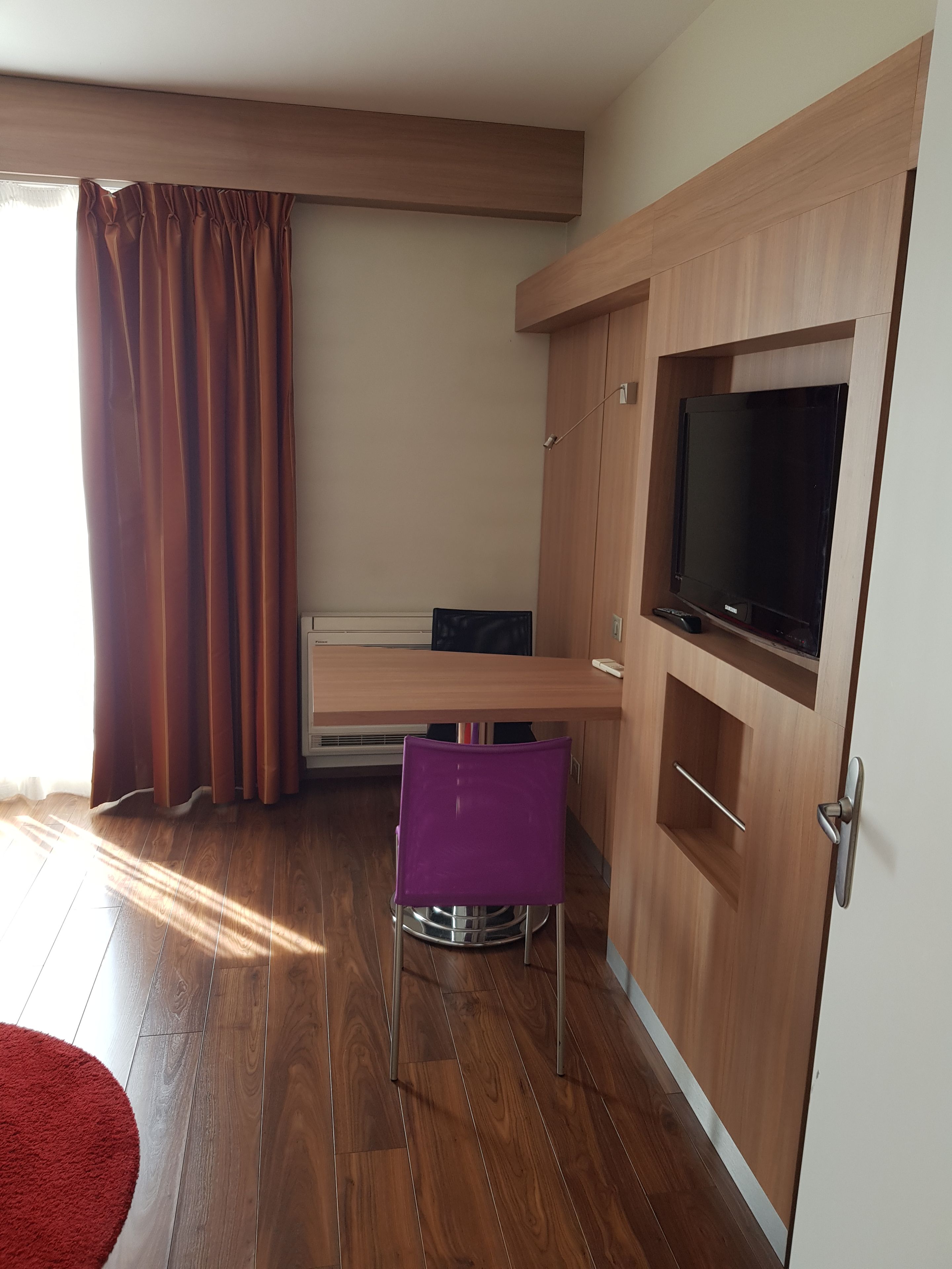 A compact room with a wooden floor, a red rug, a purple chair, a TV on a wooden cabinet, and a window with brown curtains.