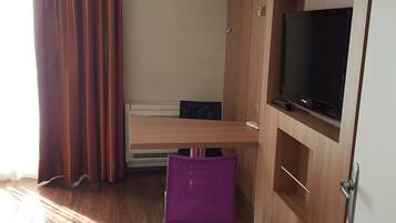 A compact room with a wooden floor, a red rug, a purple chair, a TV on a wooden cabinet, and a window with brown curtains.