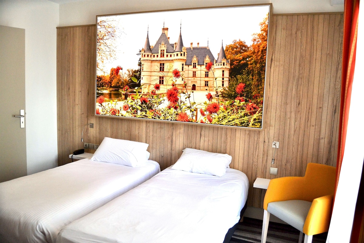 Une chambre d’hôtel avec deux lits, une tête de lit en bois, une table de chevet, une chaise et une grande fenêtre donnant sur un château.