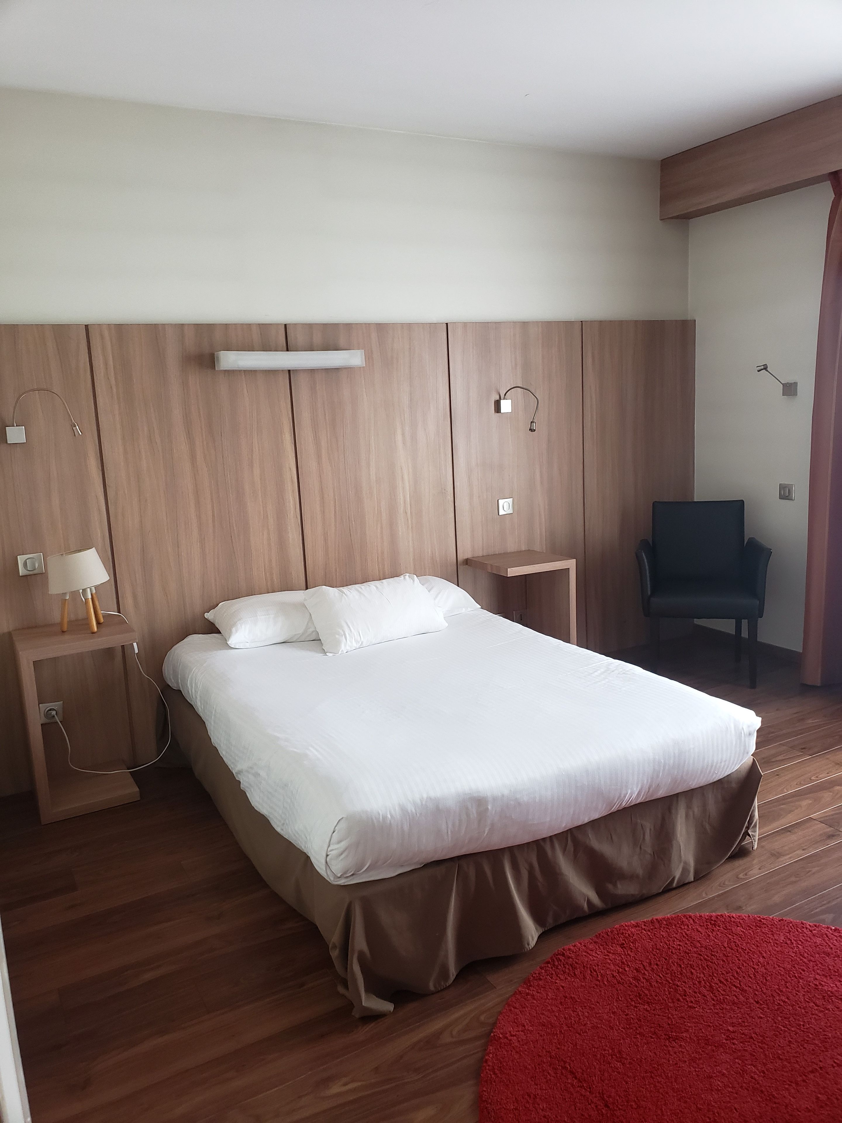 A modern hotel room with a large bed, wooden paneling, a desk with a lamp, and a red rug.