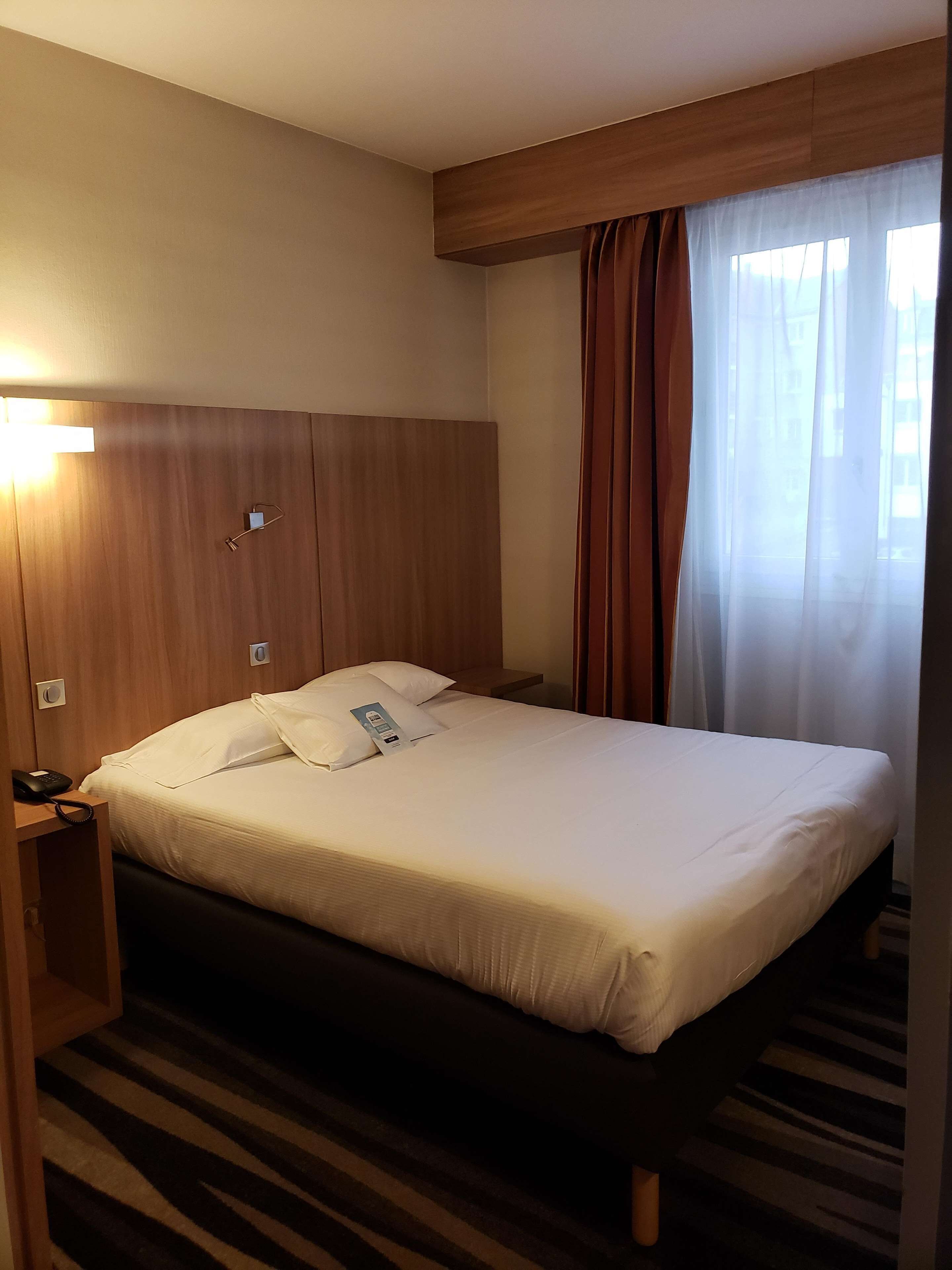 Une chambre d’hôtel avec un lit, une table de chevet avec un téléphone, une lampe fixée au mur et une fenêtre avec des rideaux.