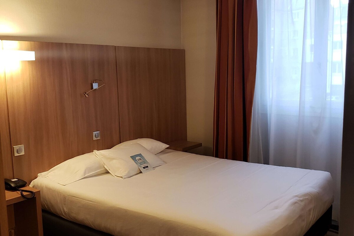 Une chambre d’hôtel avec un lit, une table de chevet avec un téléphone, une lampe fixée au mur et une fenêtre avec des rideaux.