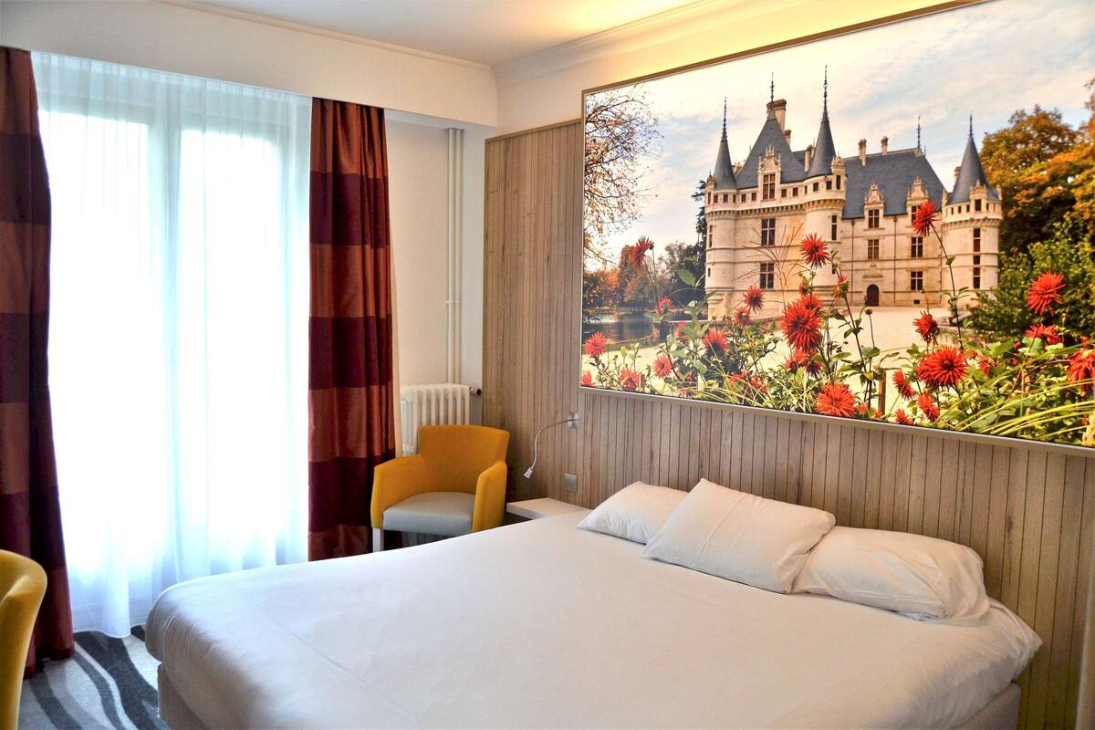 Habitación de hotel con una cama grande, una silla, una ventana con cortinas y un mural en la pared con un castillo.