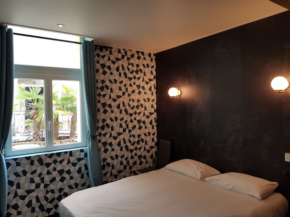 Un dormitorio con una cama, una ventana con cortinas, un papel tapiz estampado y dos luces fijadas a la pared.
