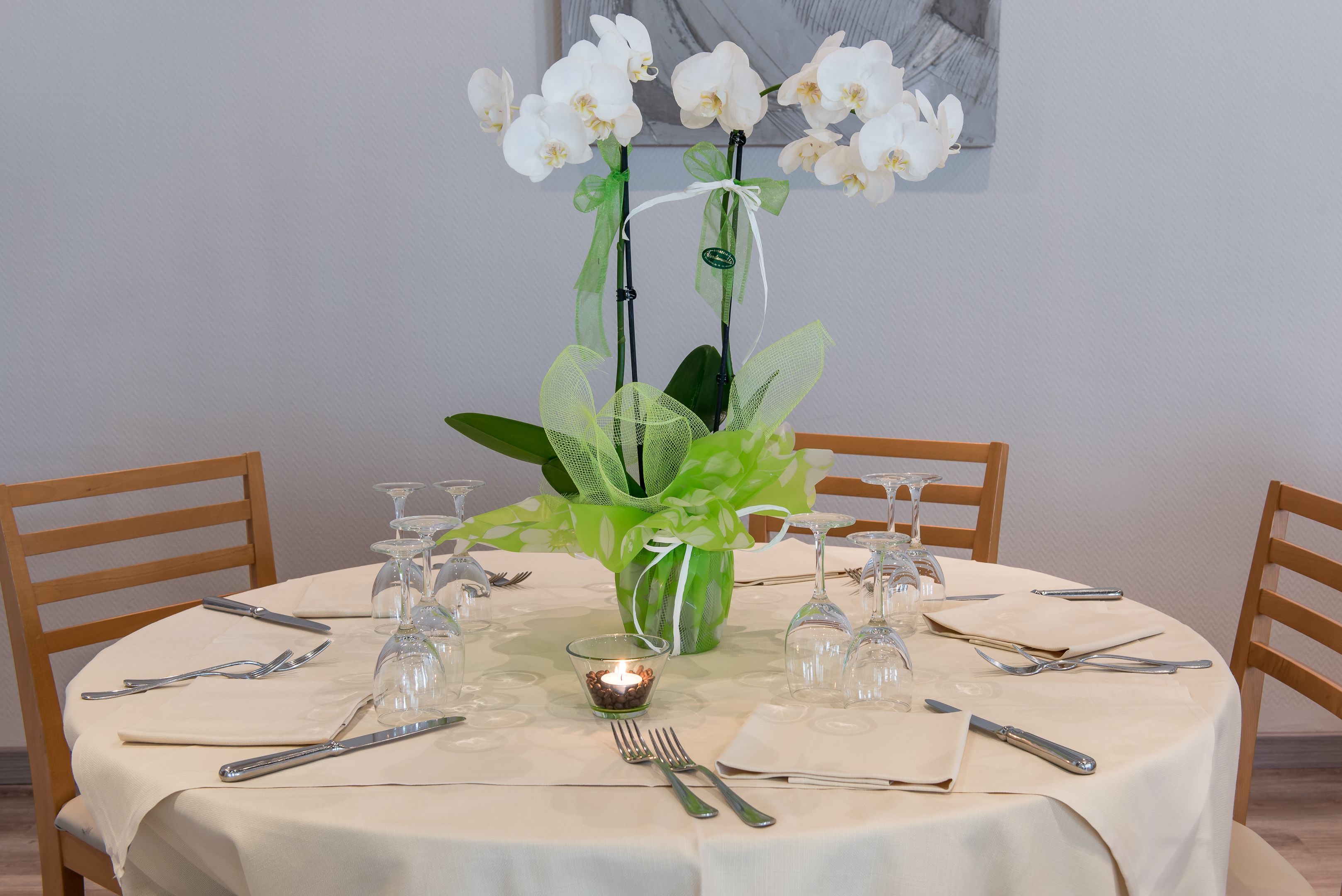 Uma mesa de jantar redonda com toalha branca, rodeada por cadeiras de madeira. No centro, um vaso com orquídeas brancas e folhas verdes.