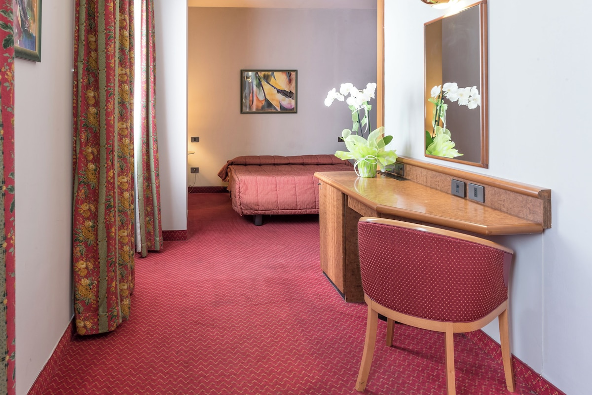 A hotel room with a red carpet, a wooden desk with a chair, a bed, a mirror, and a painting on the wall.