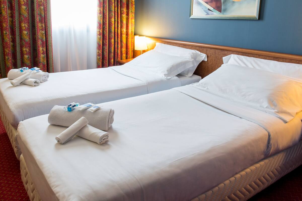 Two neatly made beds with white linens and towels, a wooden headboard, and a painting on the wall.