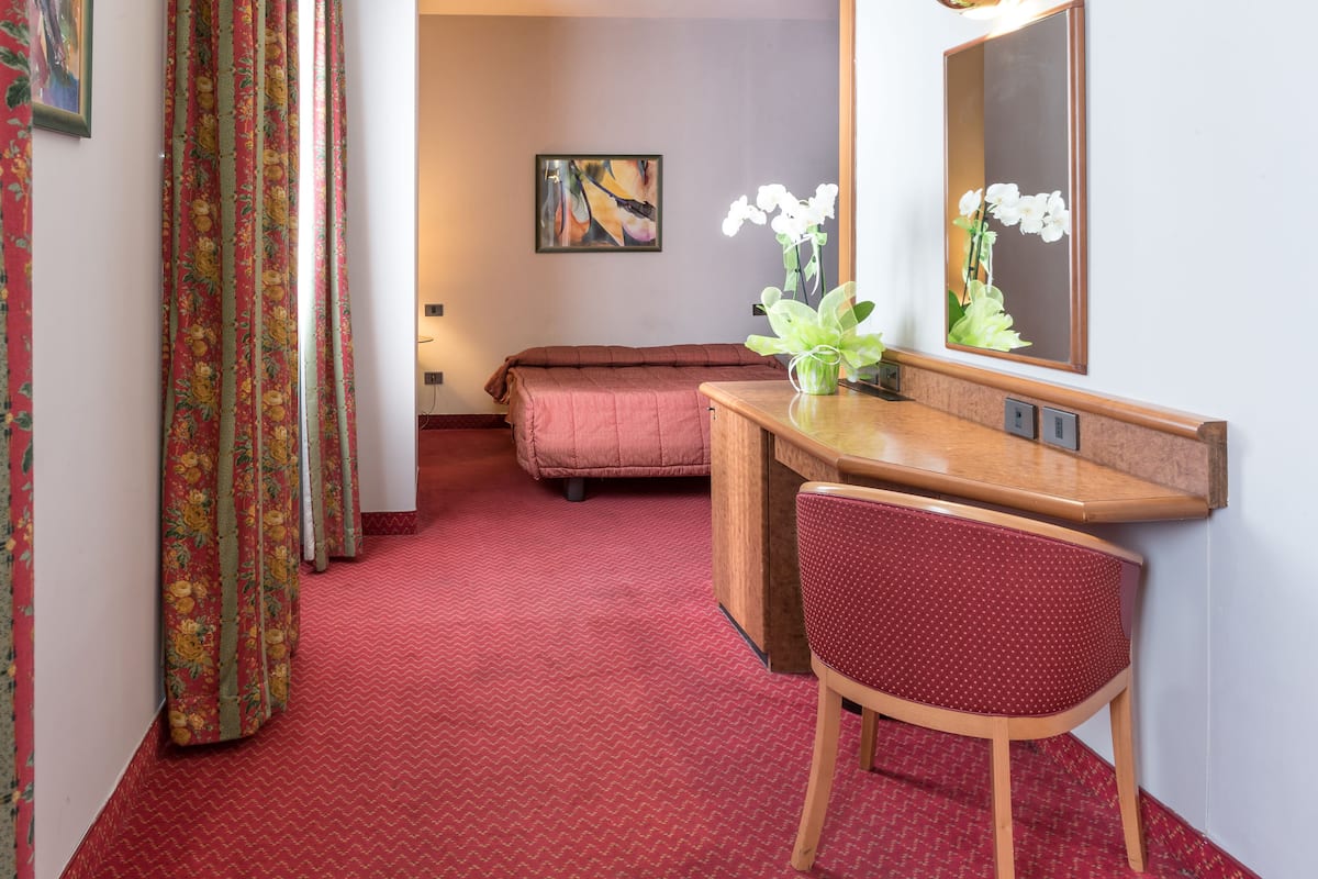 A hotel room with a red carpet, a wooden desk with a chair, a bed, a mirror, and a painting on the wall.