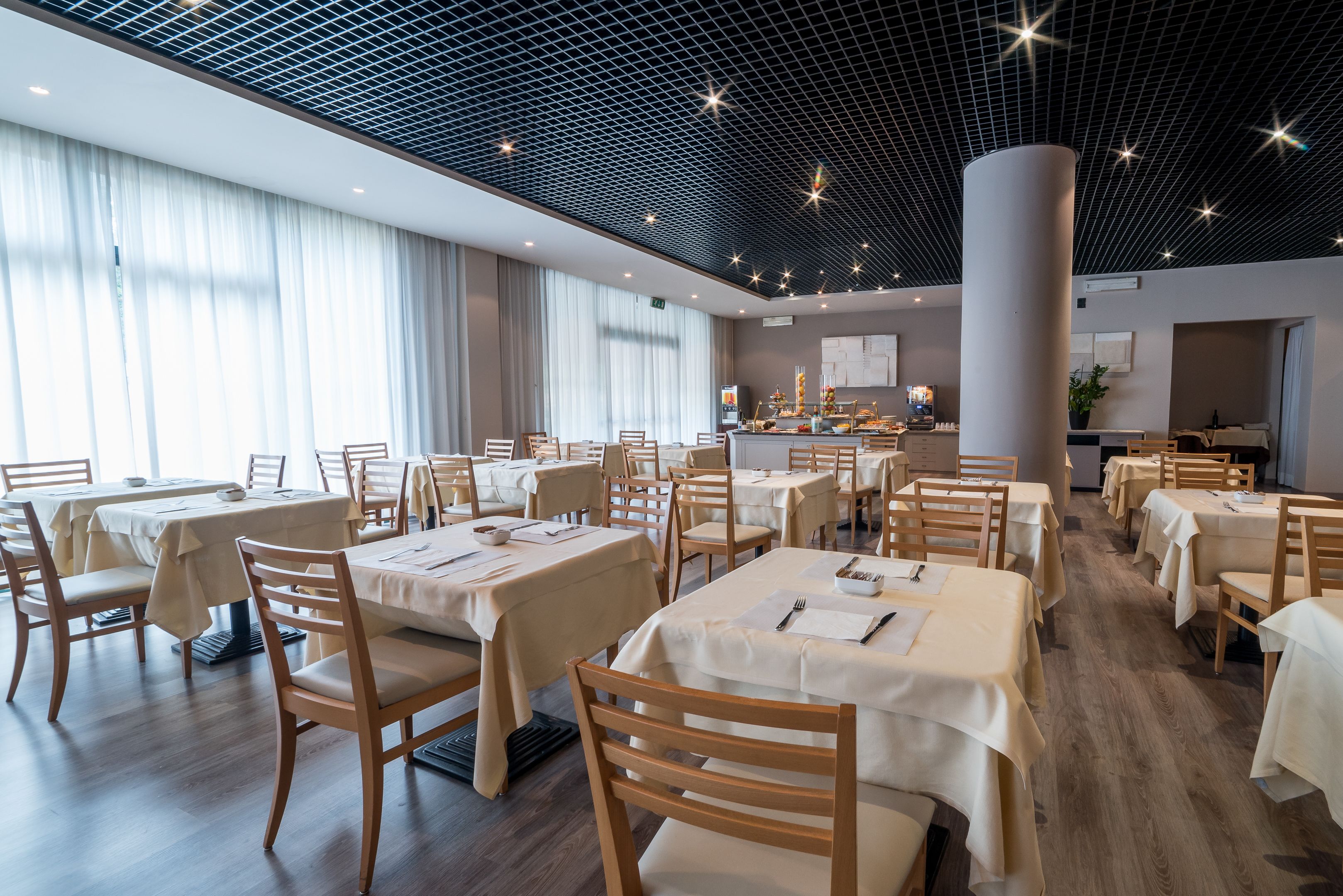 Uma sala de jantar ampla com várias mesas e cadeiras, um design de teto moderno e uma área de cozinha visível ao fundo.