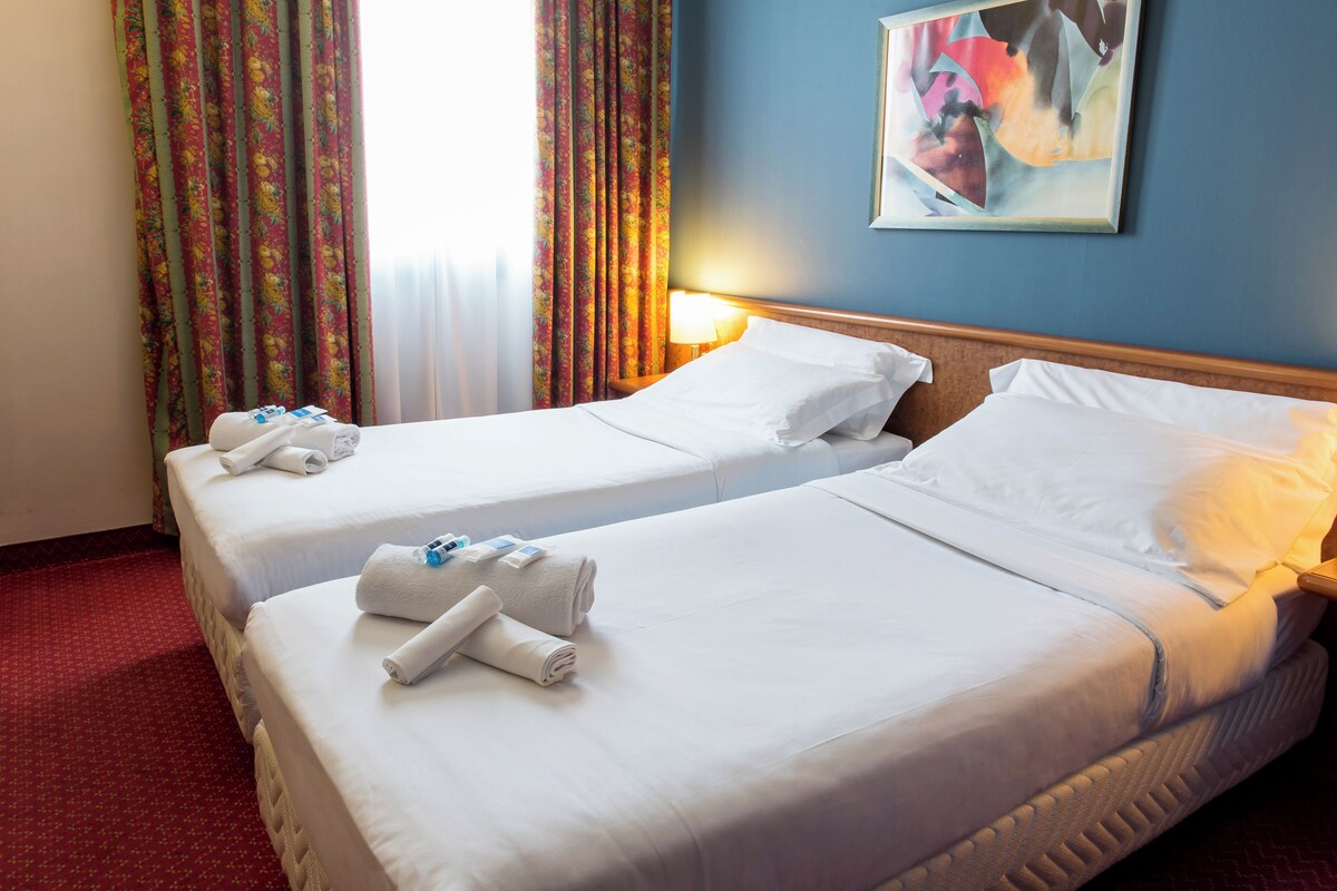 A hotel room with two beds, each with white linens and towels, a nightstand with a lamp, and a painting on the wall.