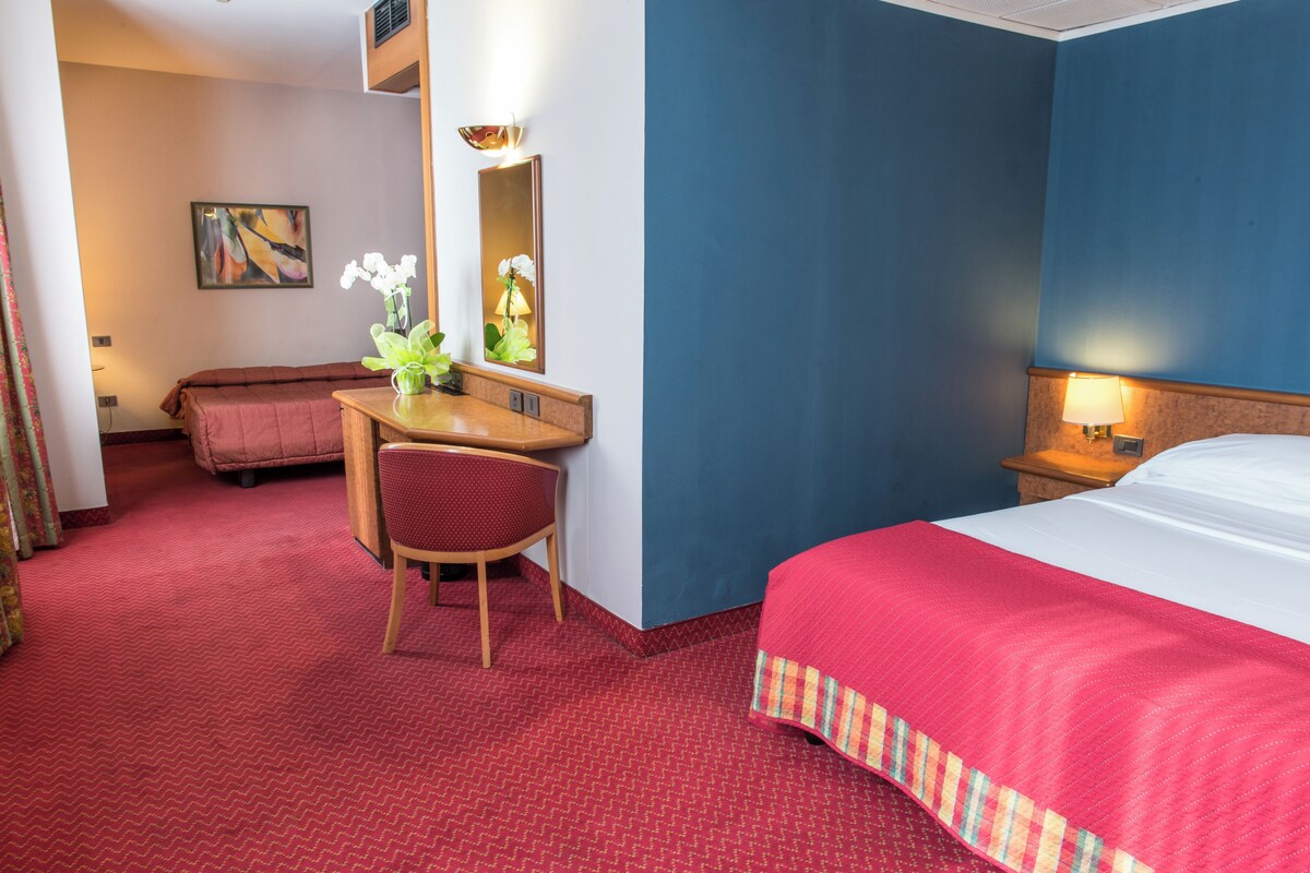 A hotel room with a bed, a desk with a chair, a small table with a vase of flowers, a mirror, and a painting on the wall.