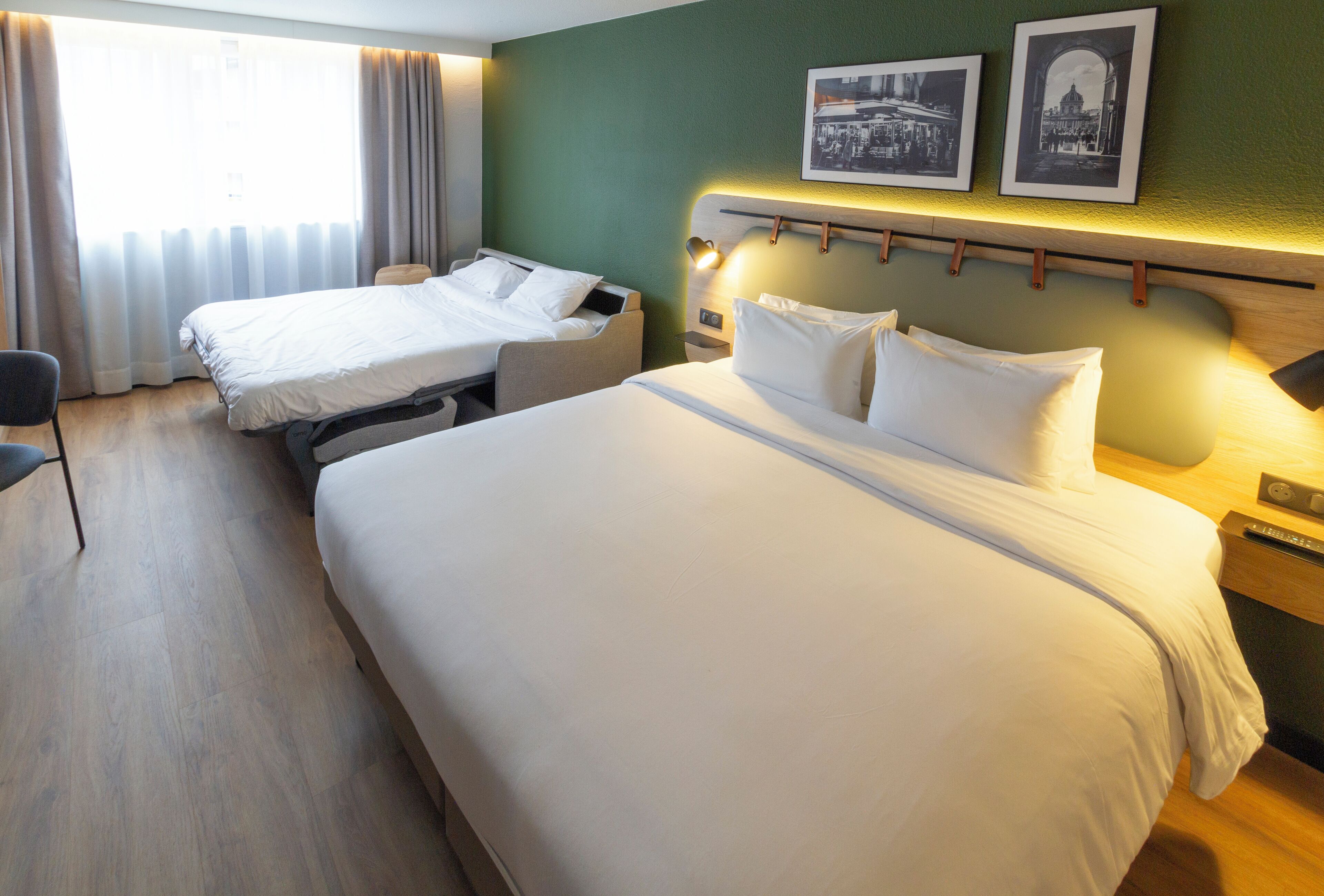 A hotel room with two single beds, a desk, and a chair. There are two framed pictures on the wall above the beds.