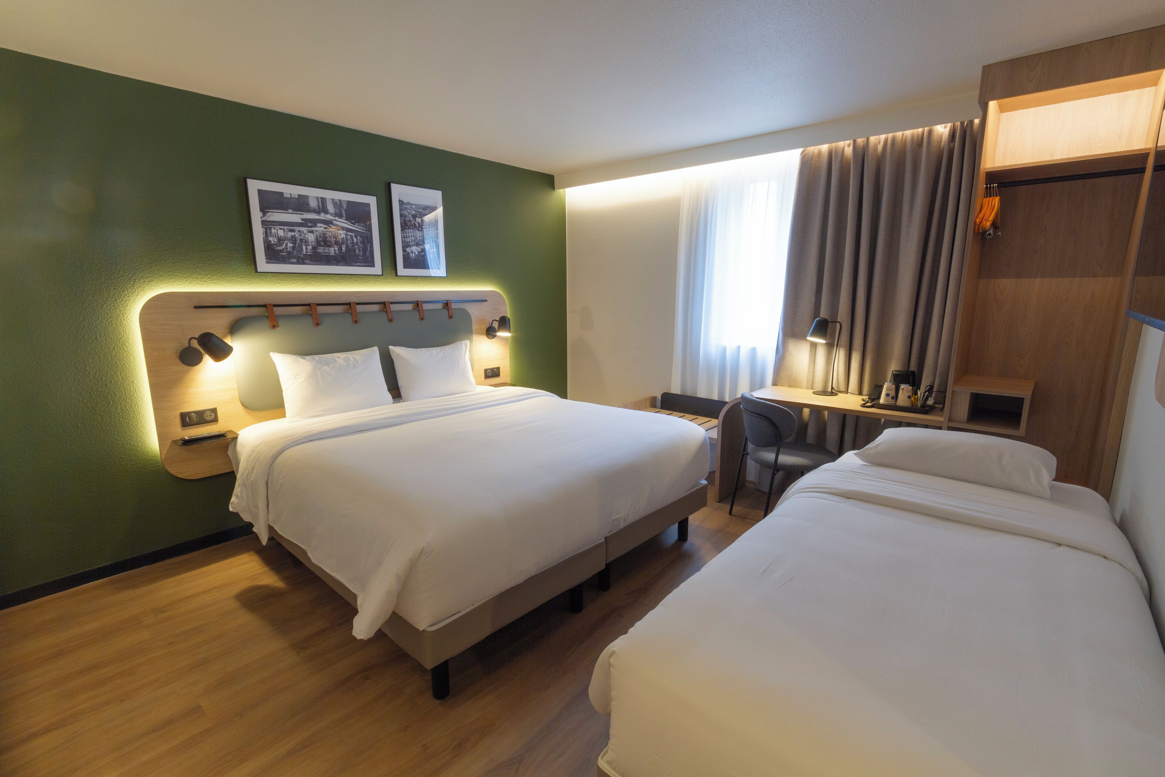 Habitación de hotel con dos camas, un escritorio y una silla. Hay dos cuadros en la pared, una ventana con cortinas y un armario de madera.